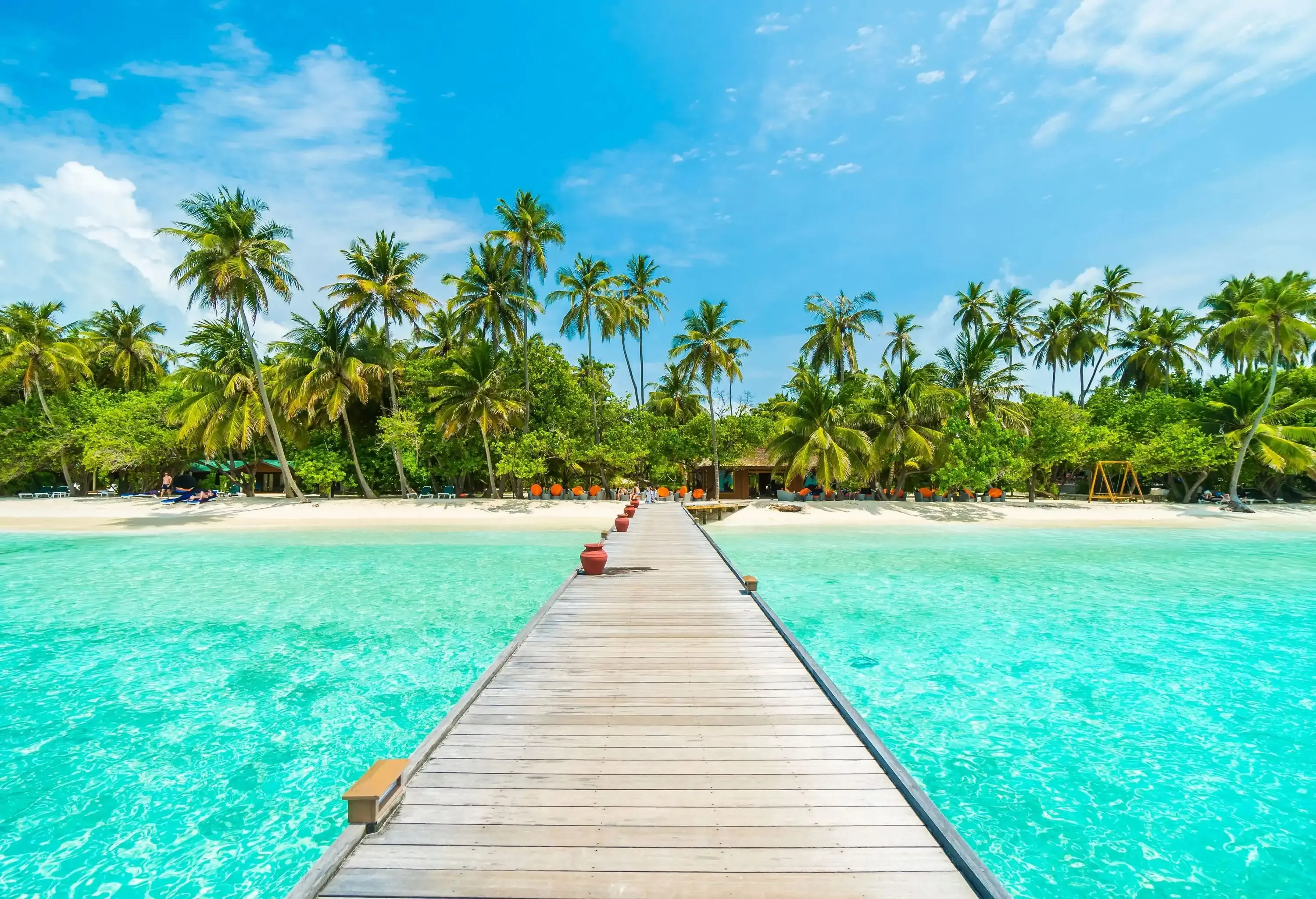 A wooden dock running across the turquoise sea waters, leading to a beach surrounded by tropical greenery. 