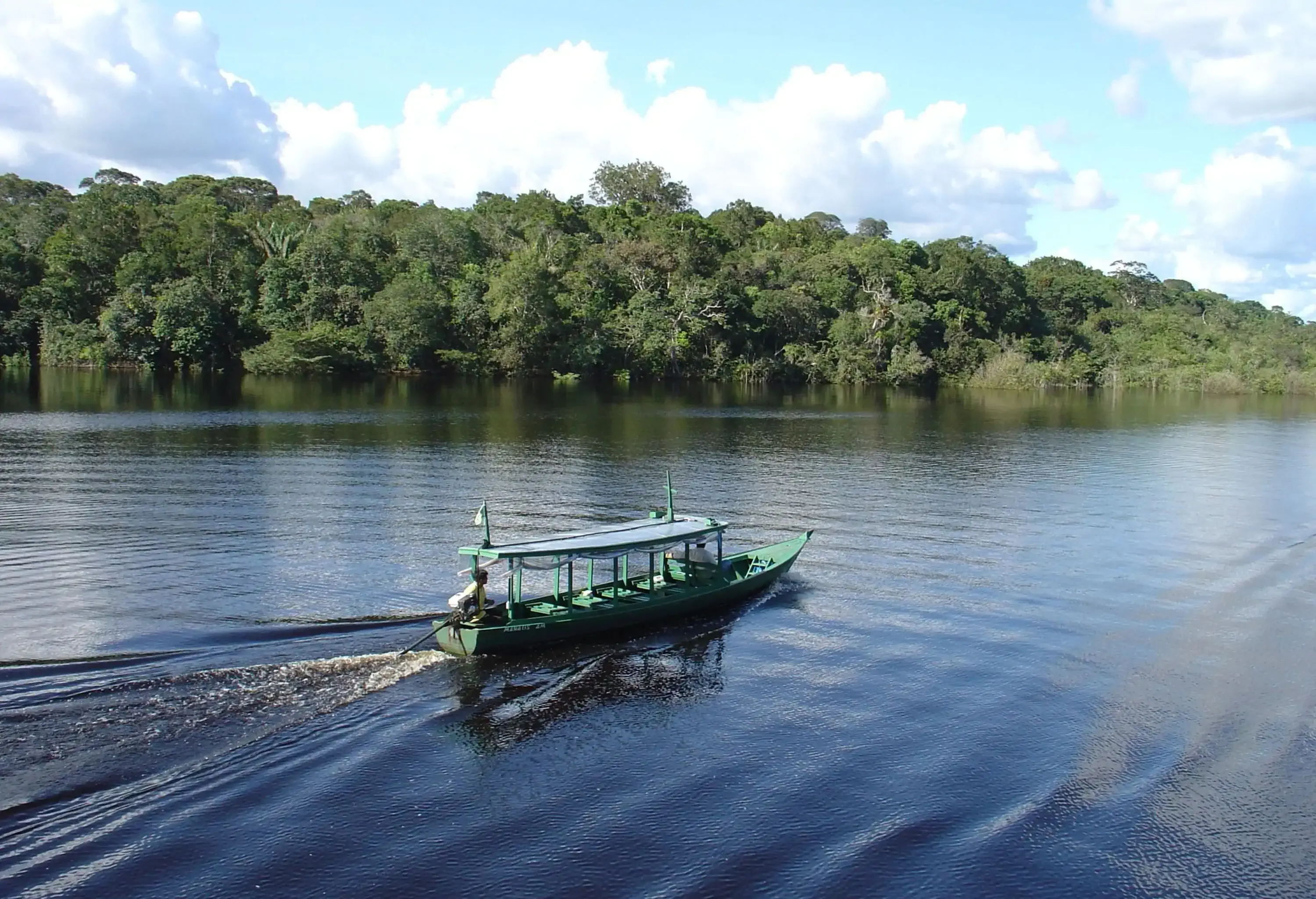 A boat slowly travelling down the river, with a lush forest along the banks in the background.