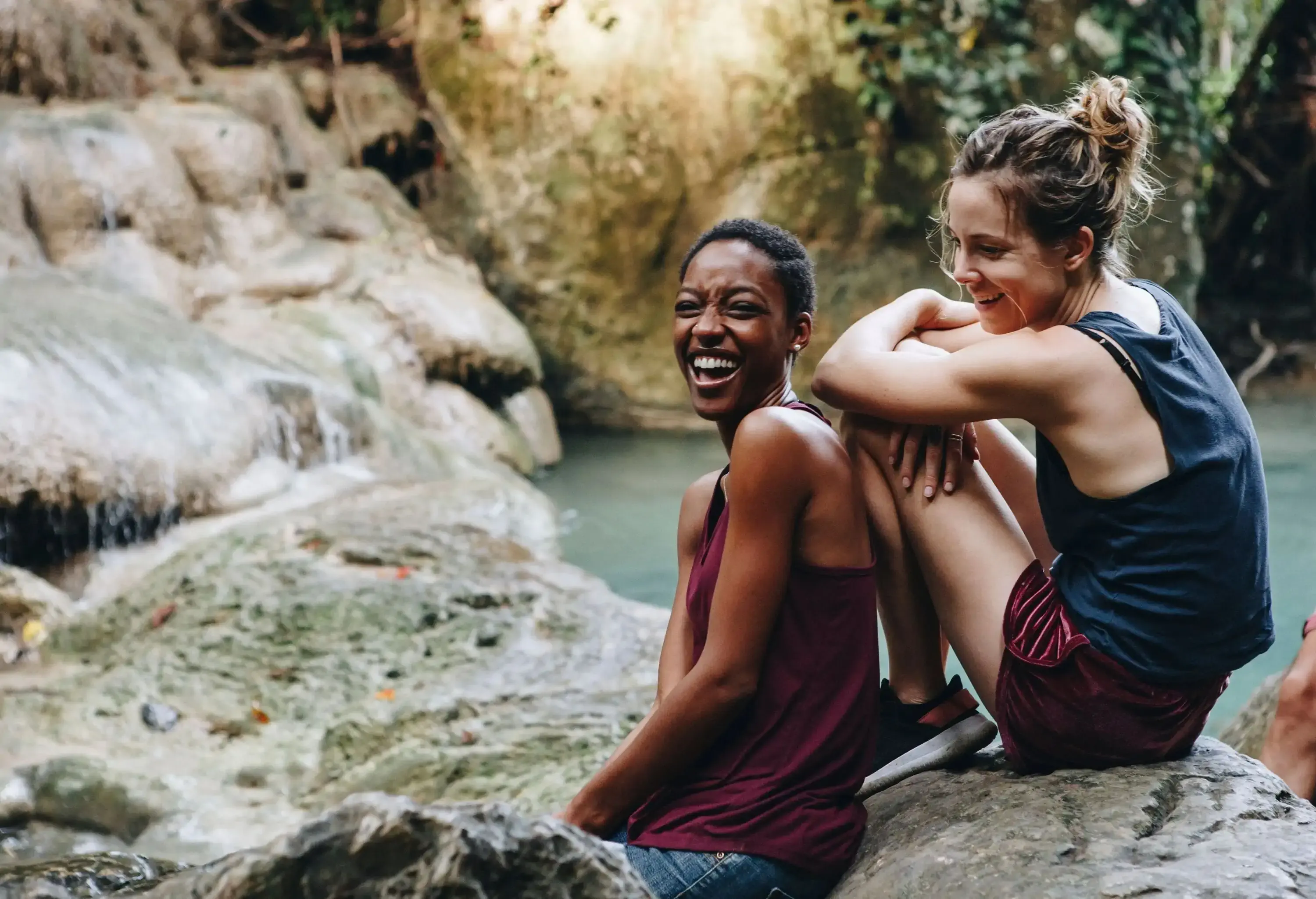 Two friends hang out along a rocky stream, one sitting on a boulder and the other leaning on it.
