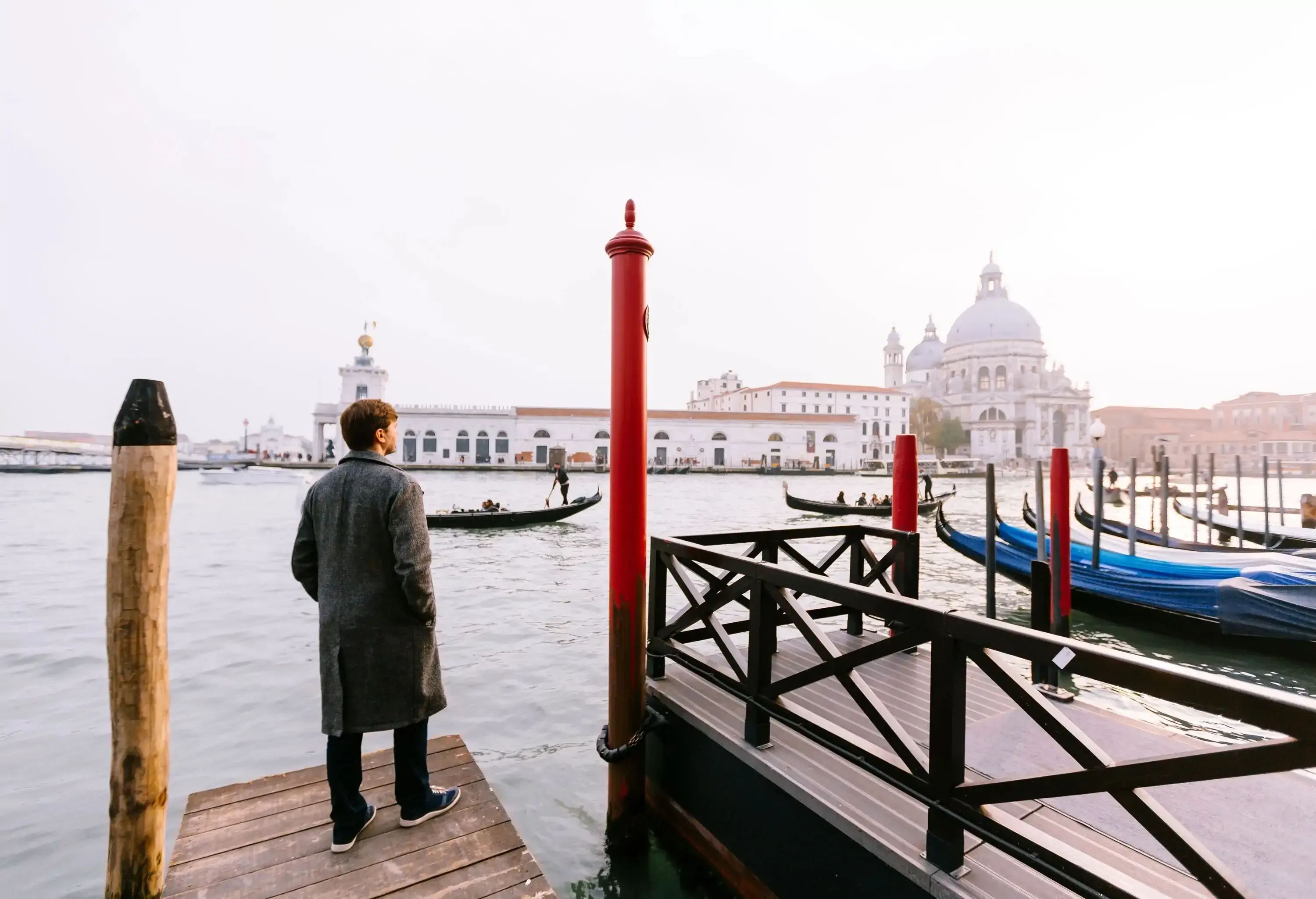 A person donning a long coat stands pensively on the pier, gazing intently at a majestic white building adorned with a grand dome across the shimmering waters.