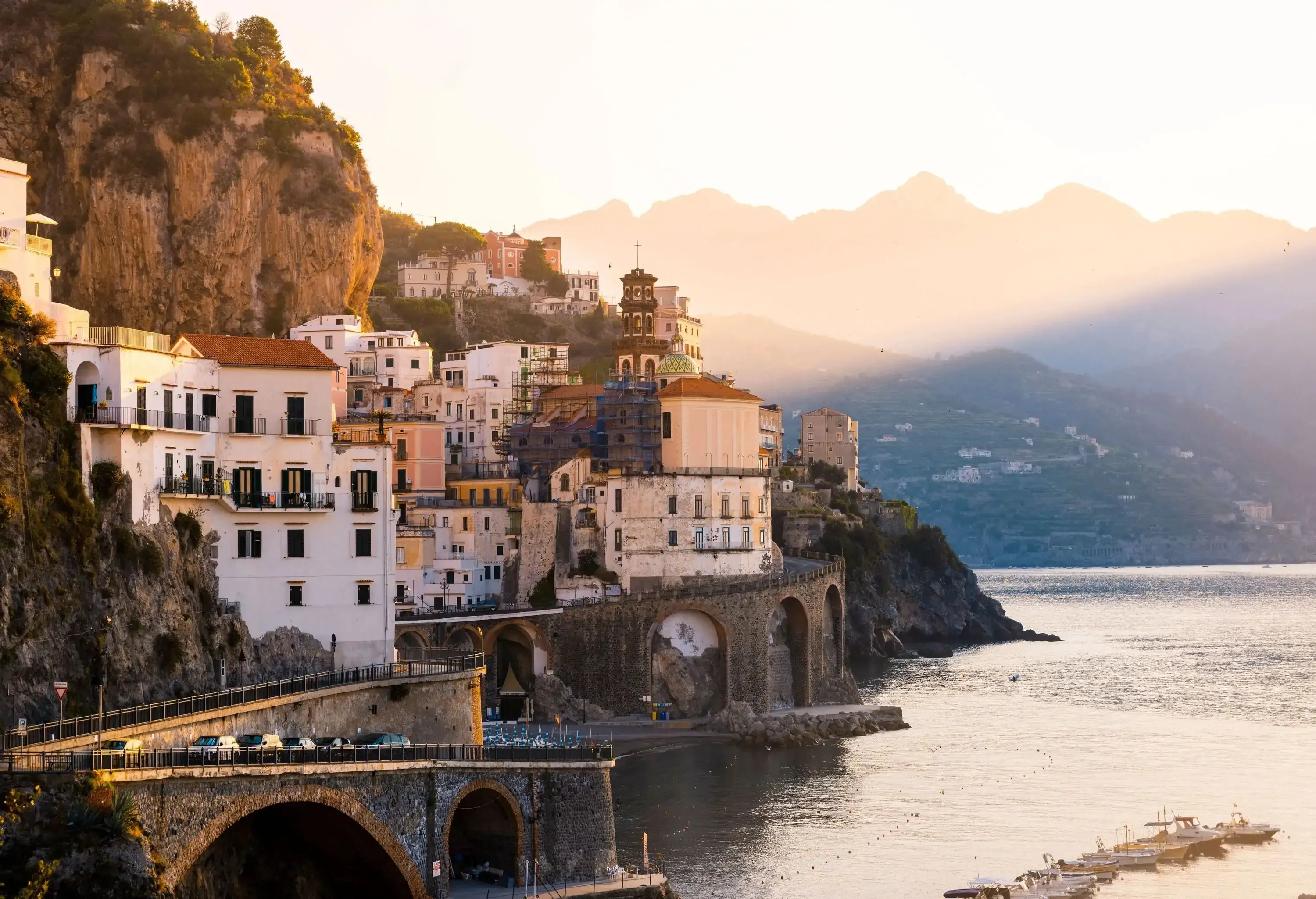 A sunlit coastal village on the steep mountainside overlooking the lined small boats on the sea.