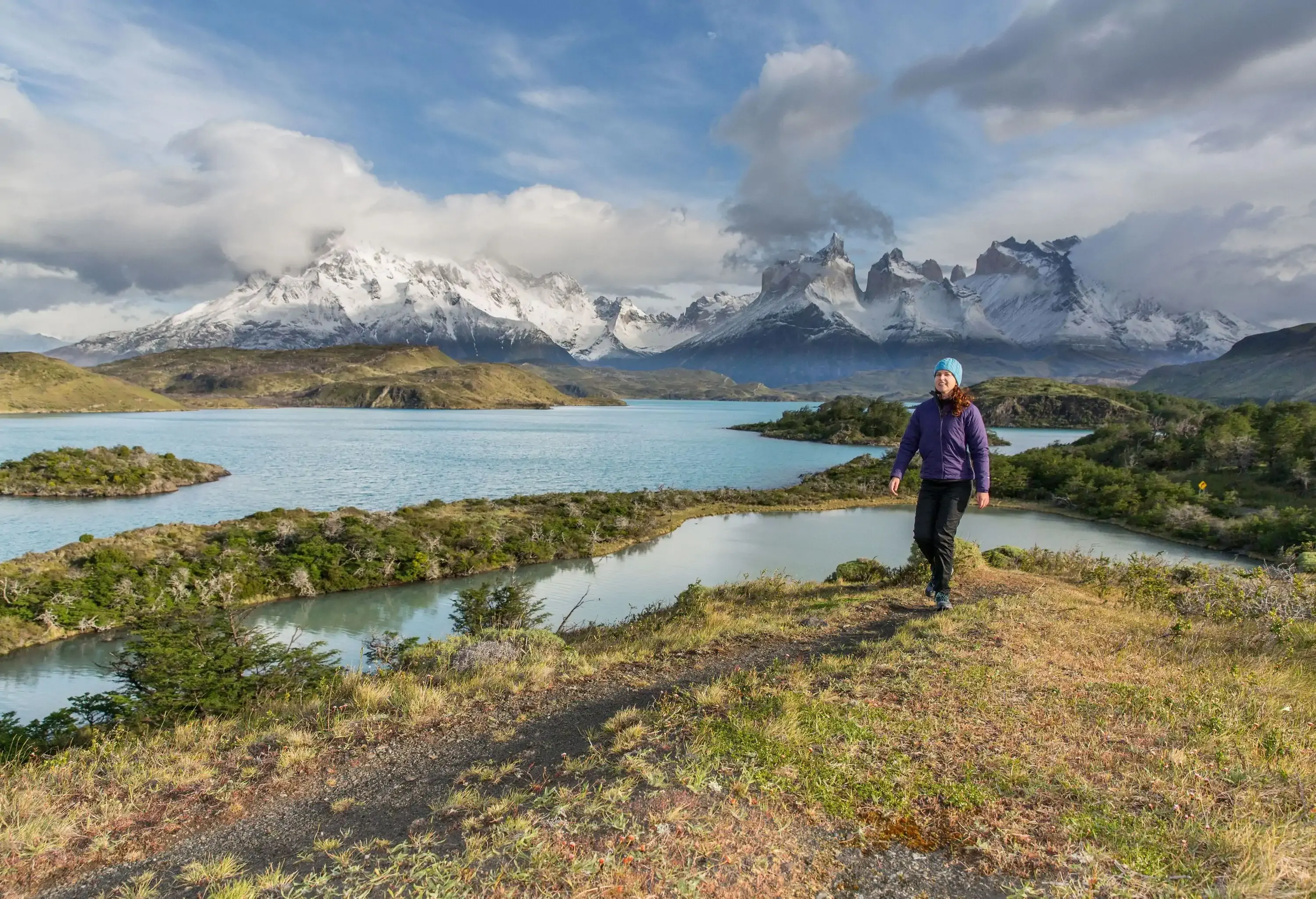 A woman wearing a blue bonnet cap trotting down a lakeside trail, with rugged snow-covered mountains in the backdrop.