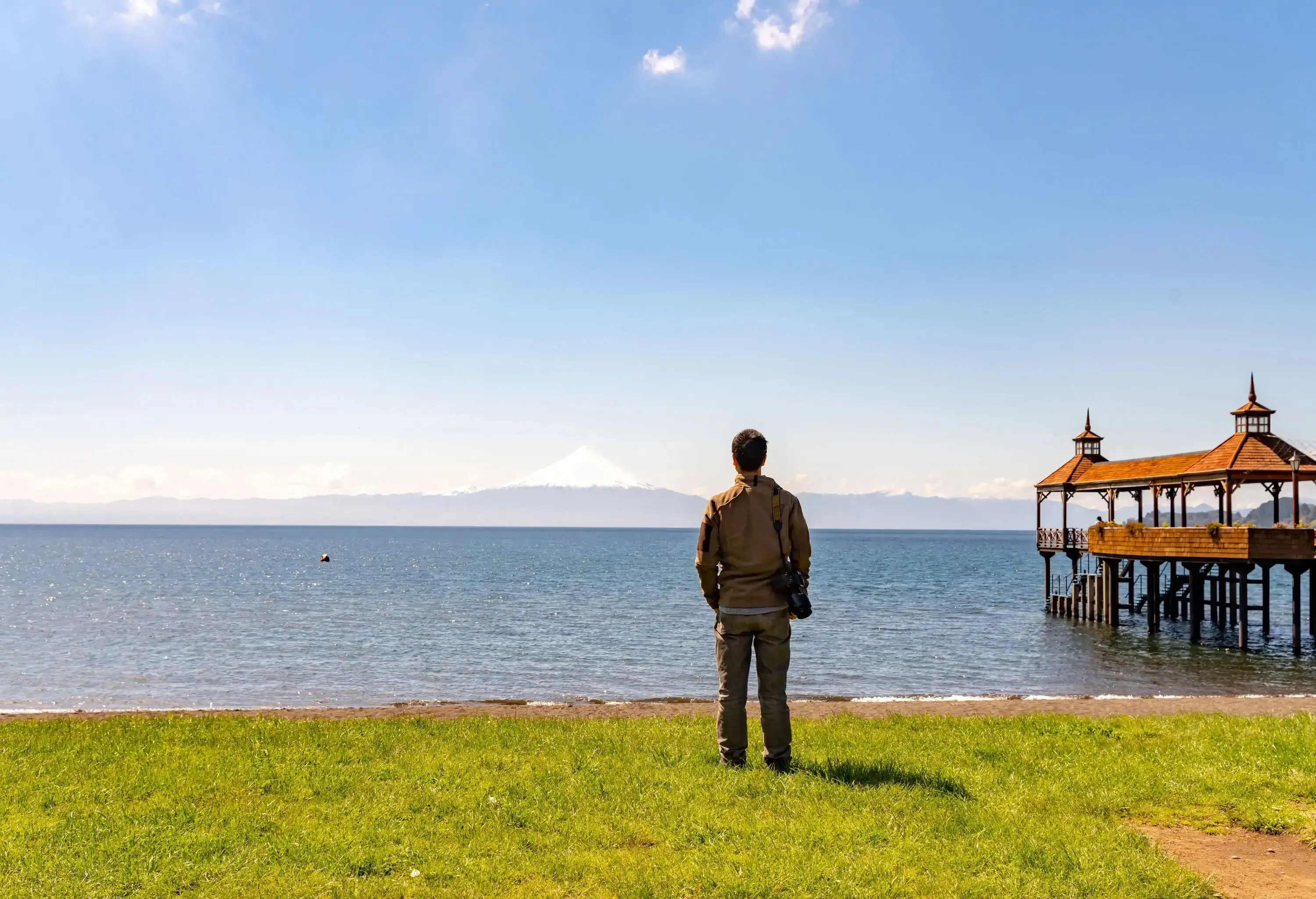 A man with a digital camera slung over his right shoulder standing on a grassy beach, facing the sea.