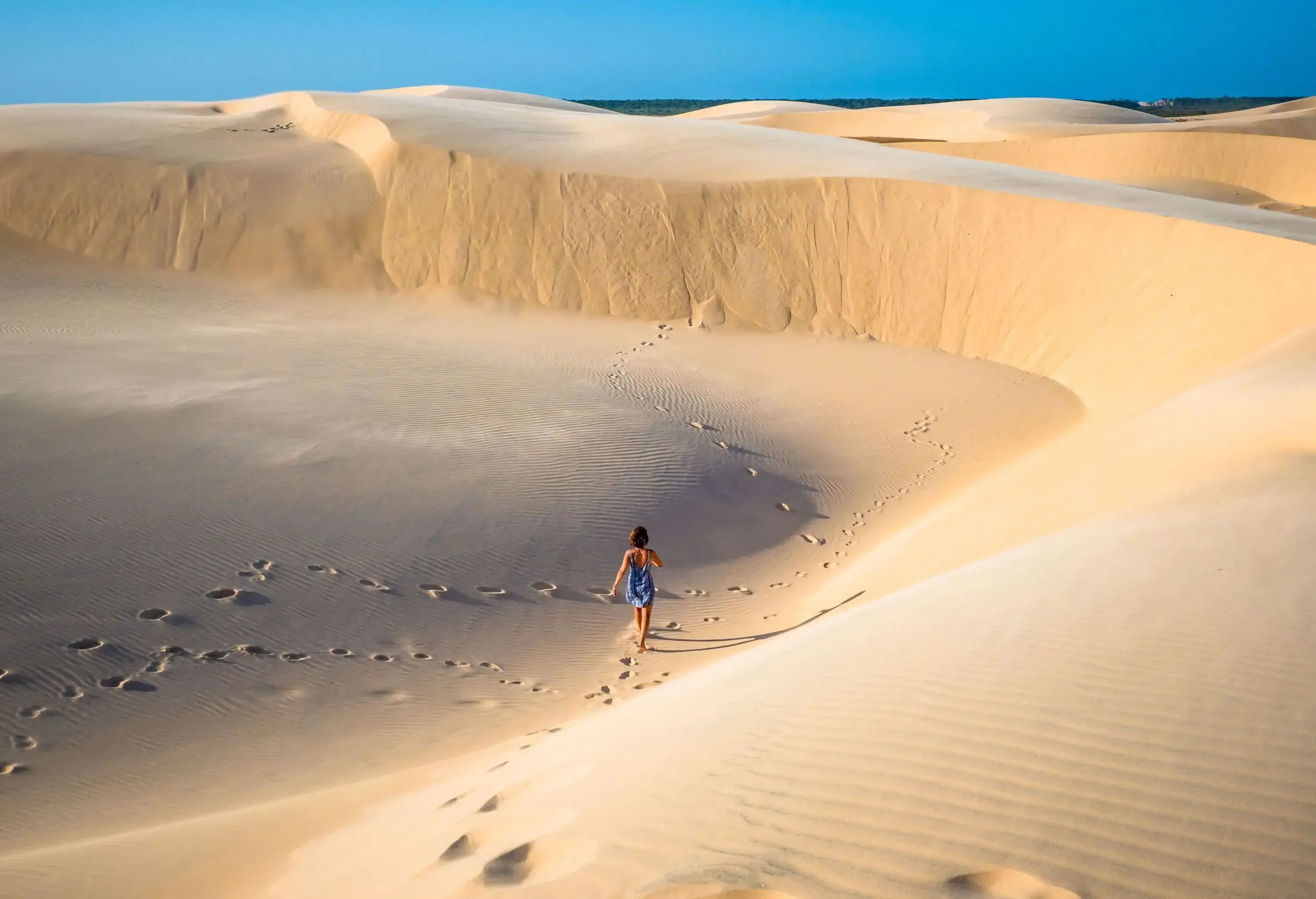 A woman wandering across a fine sand dune with traces of footprints behind her.