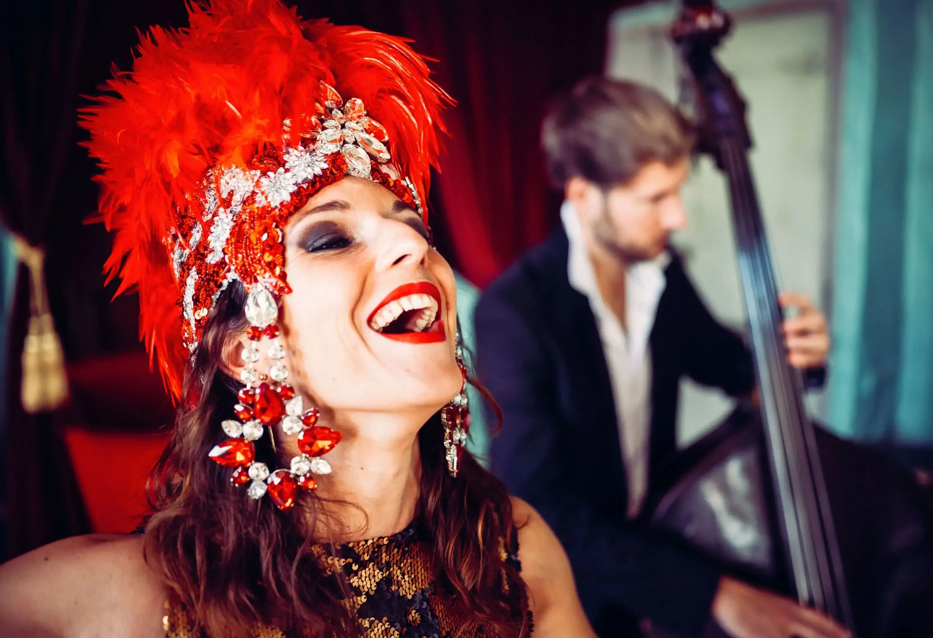 A person wearing a crimson feathered headdress with crystals set in a flowery pattern smiles widely.