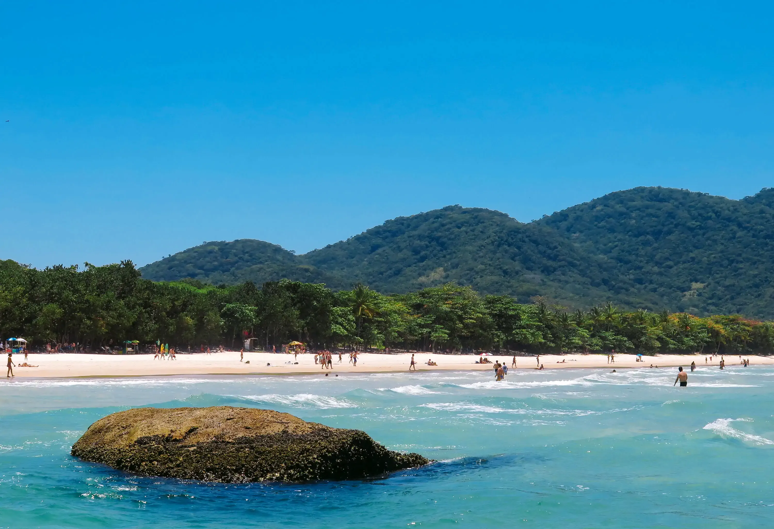 A rugged rock juts out from the water in the foreground as beachgoers enjoy the idyllic scene, with a picturesque beach lined with tropical trees stretching out in the middle ground and verdant hills providing a stunning backdrop in the distance.