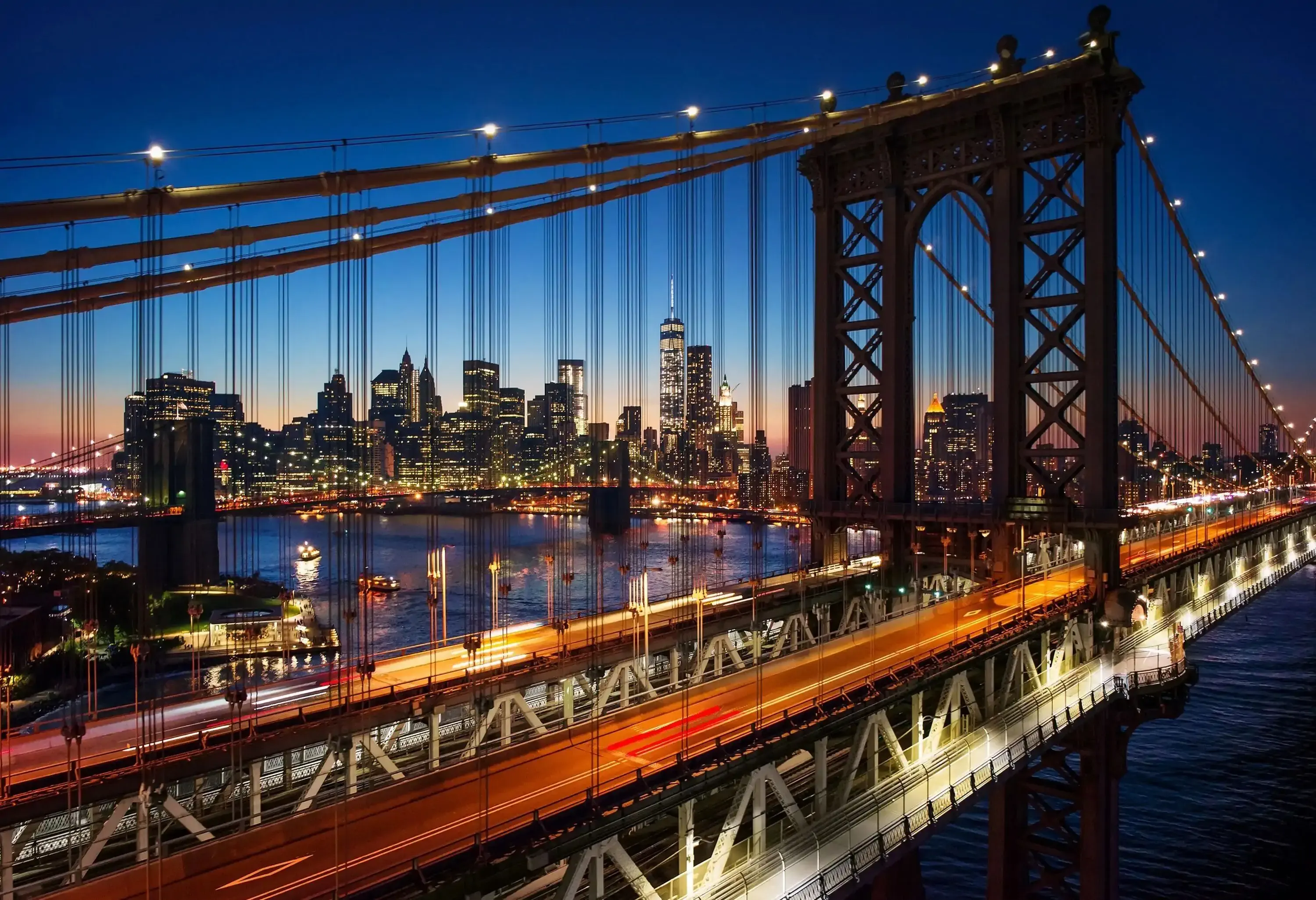 A night-time view of a busy and bright road bridge across a calm bay with views of a dazzling city skyline in the distance.
