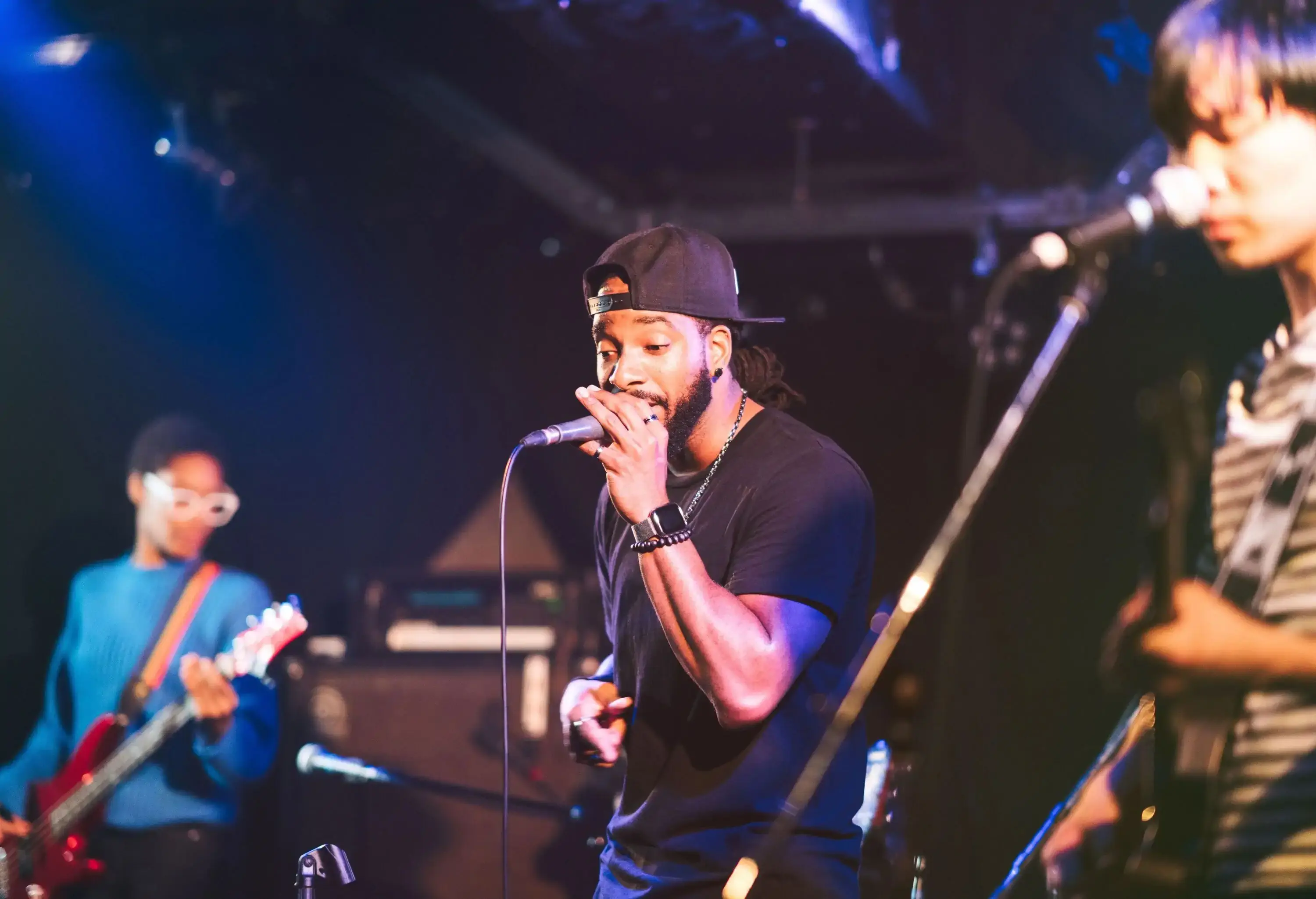 A multi-ethnic music group electrifies the stage, with a vocalist in a reverse cap gripping the microphone, passionately singing and captivating the audience.