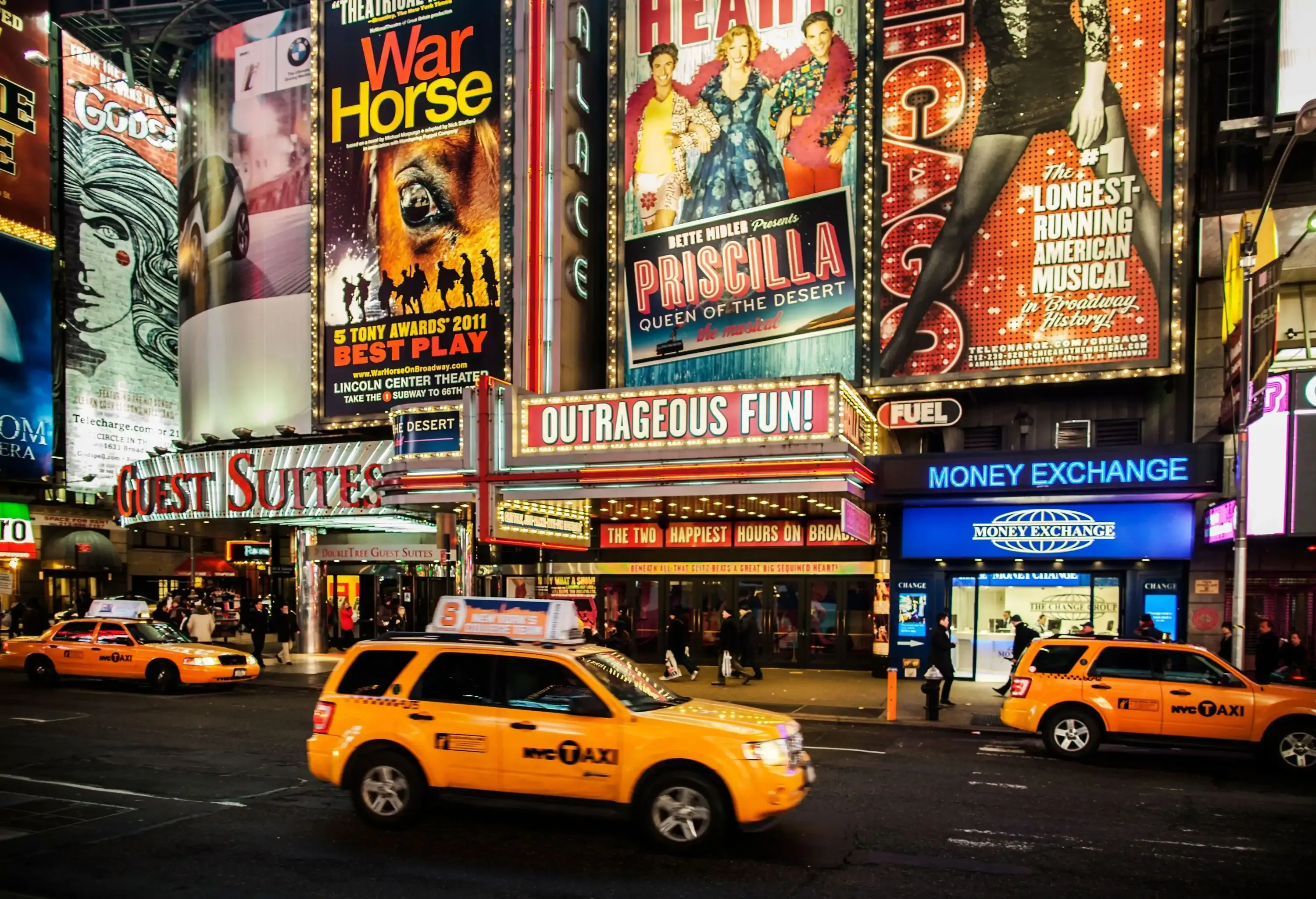 Iconic Broadway theatres in the heart of Times Square, New York City, come alive with dazzling poster ads, as yellow taxis in the foreground add a vibrant touch to the bustling scene.