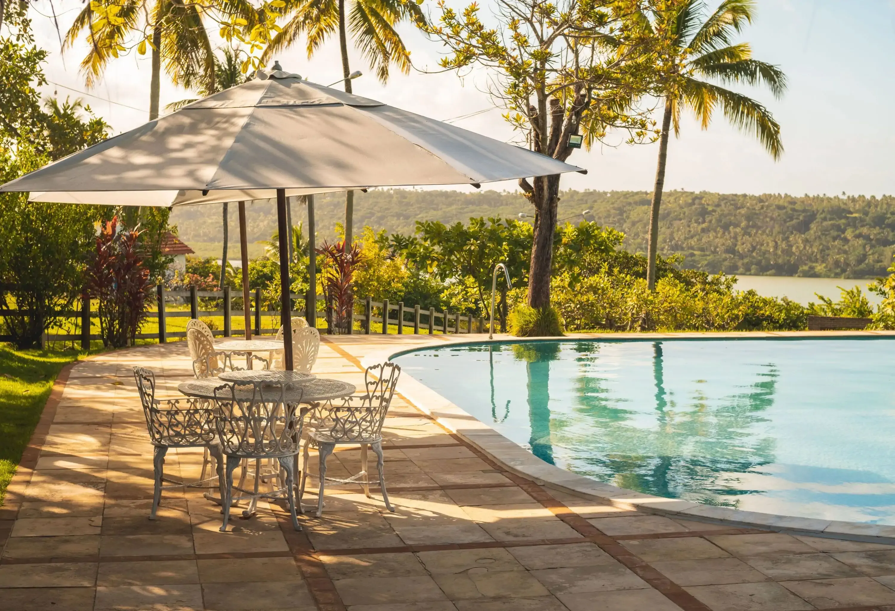 An outdoor seating area under a big umbrella beside a pool with views of tropical trees and forested mountains.