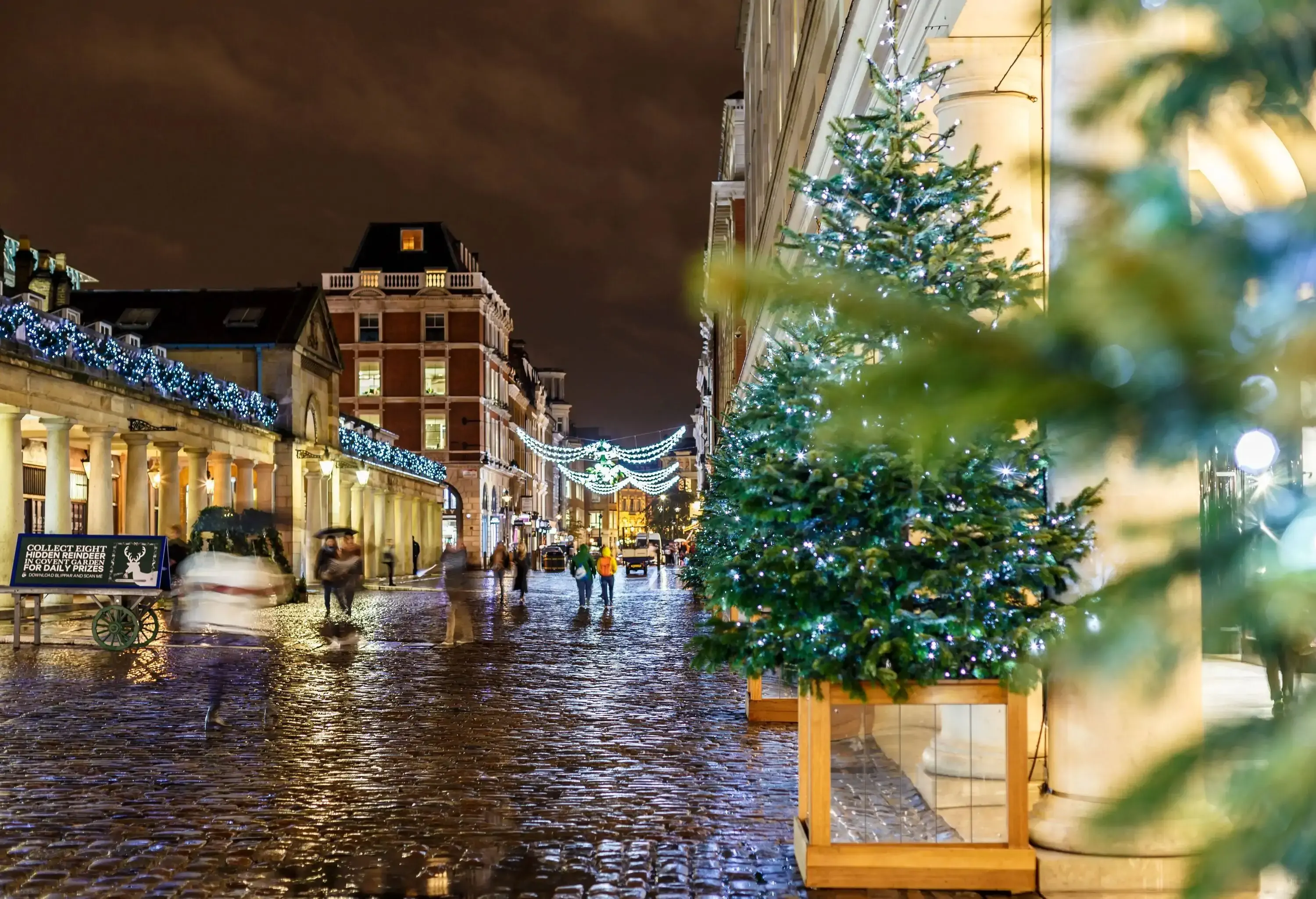People are in motion in the cobbled stone street lined with classic buildings and brightly lit by Christmas lights.