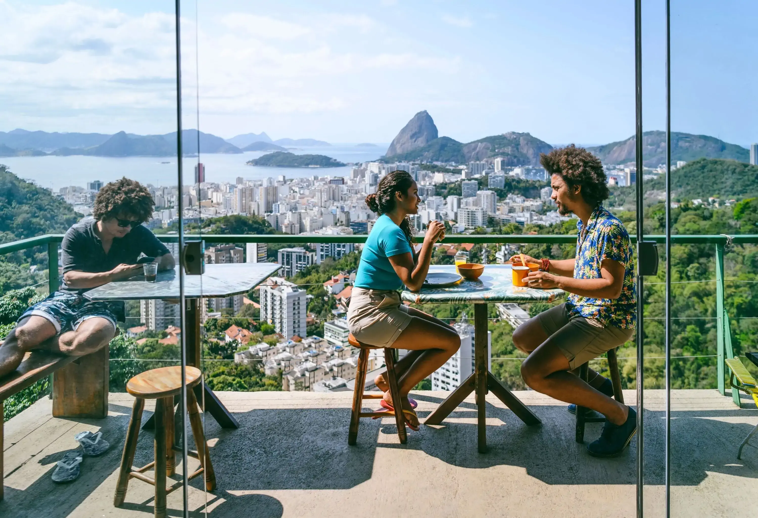 A pair and a man sitting at two separate tables on a terrace with views of the city and its natural surroundings.