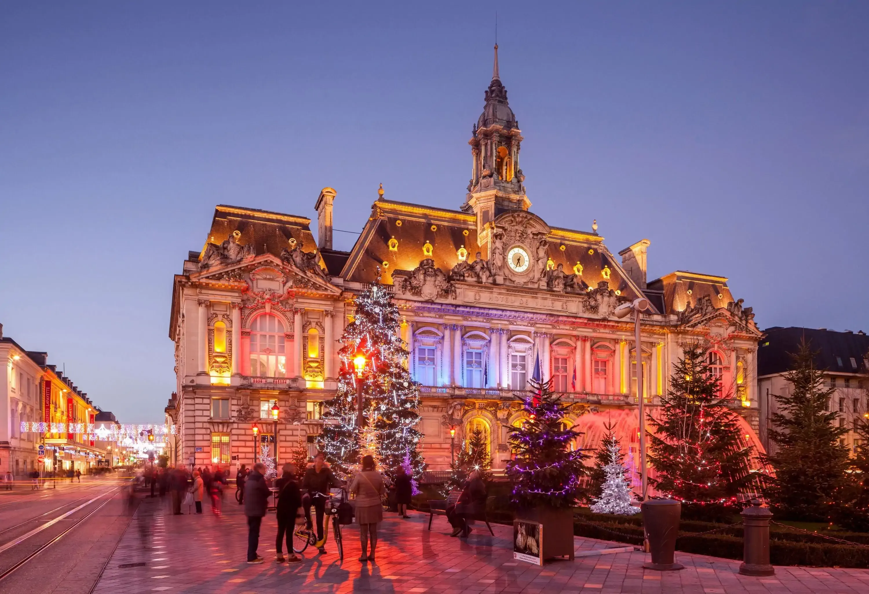 An exquisite Renaissance-style building shining in different colours with numerous illuminated Christmas trees in its front yard.