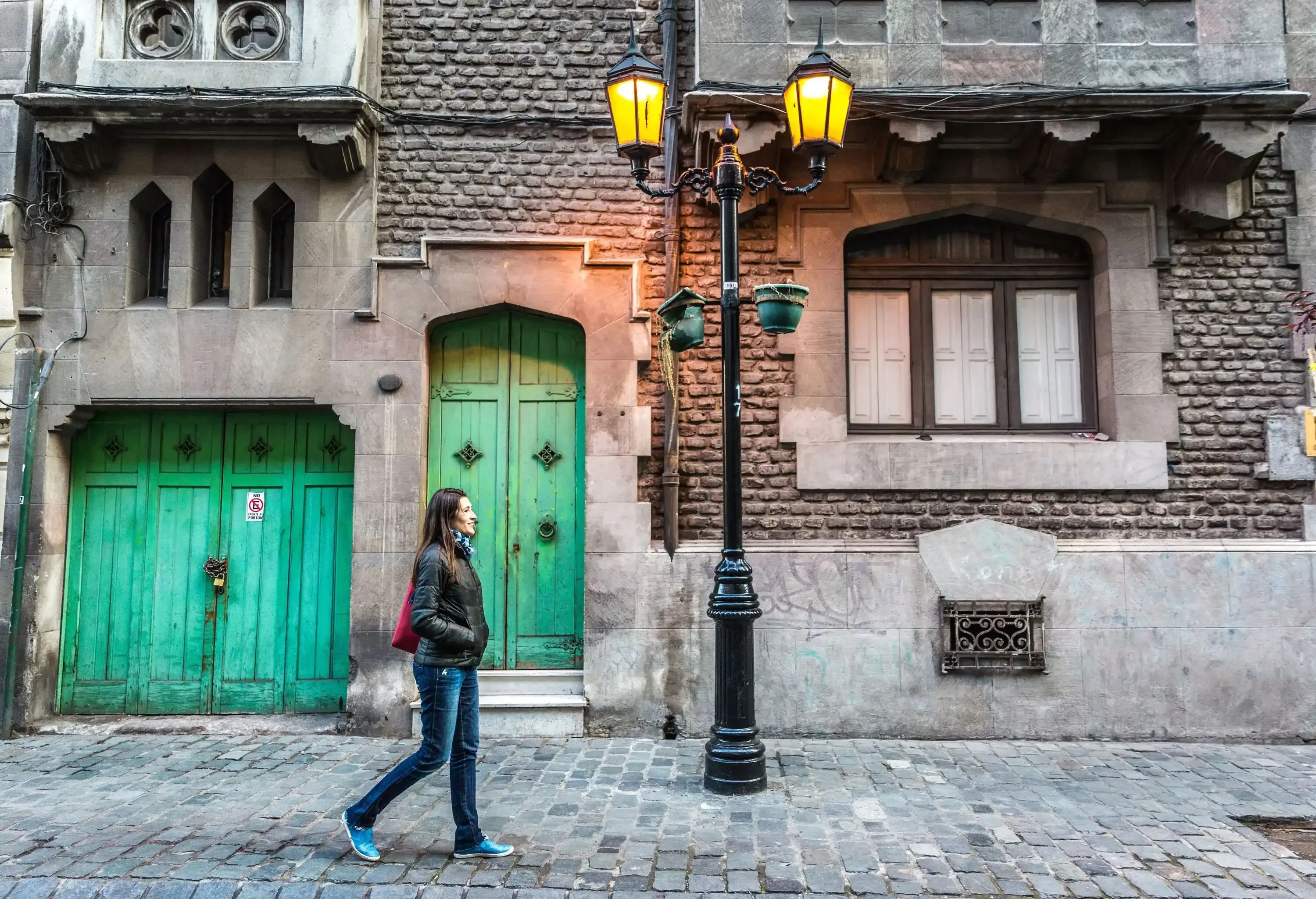A woman walking on a pavement with a light post next to a stone building with green doors.