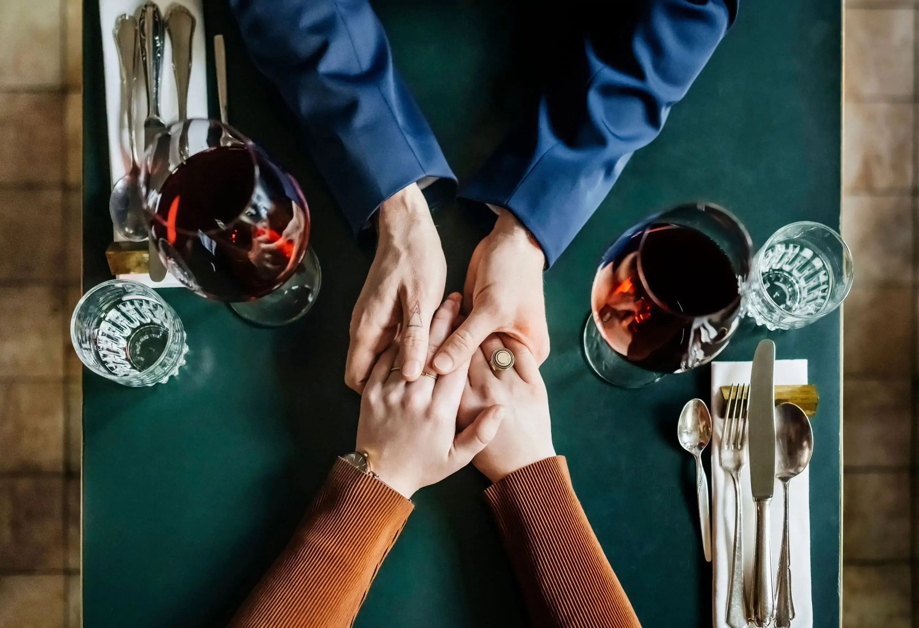 A romantic couple holds hands across a table set with gleaming utensils and wine and water glasses.