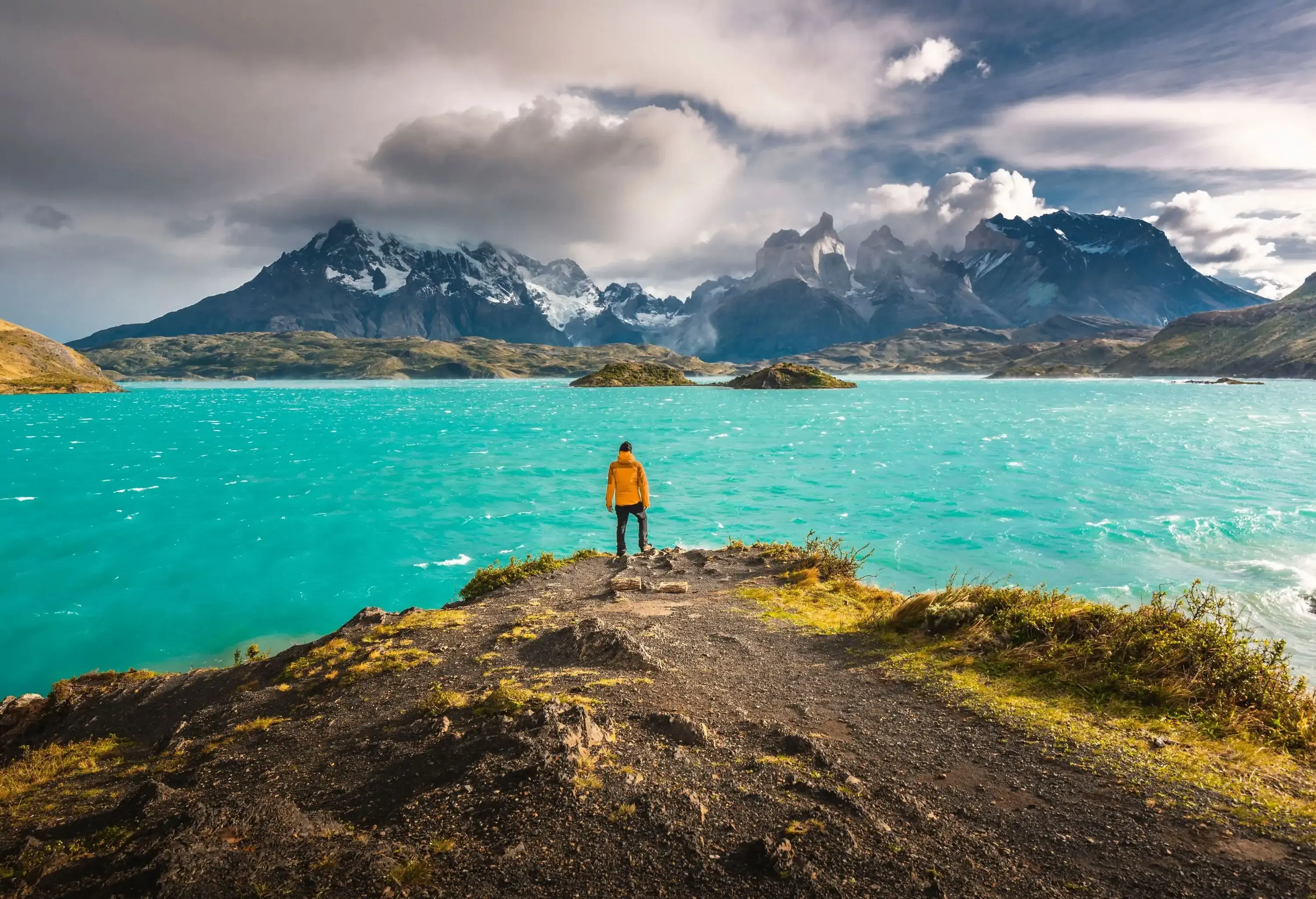 A man in a yellow jacket perched on a headland looking over a stunning lake and mountains in the distance.