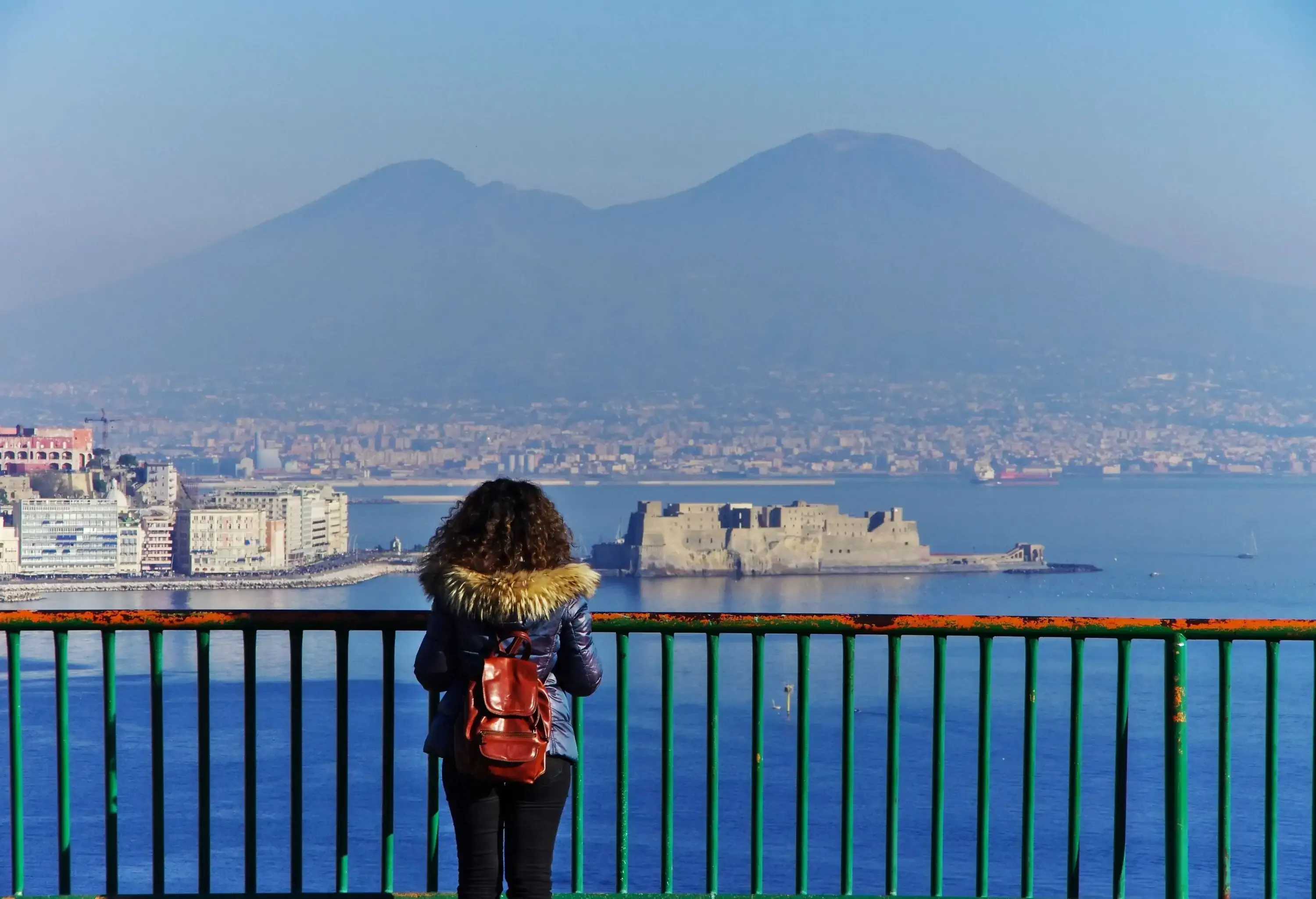A woman wearing a parka stand on balcony while gazing over the seafront castle and cityscape.