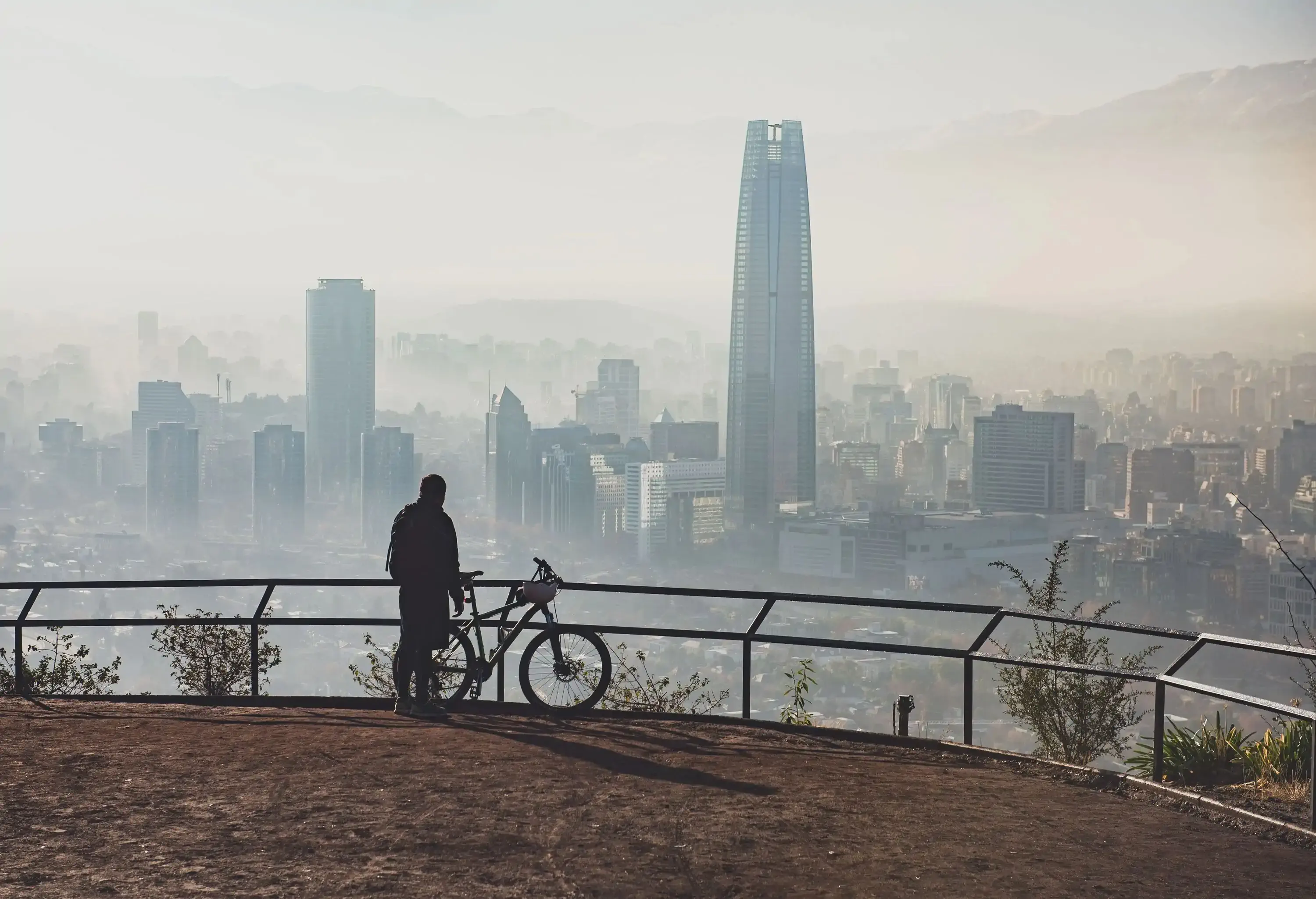 A man with a bicycle stands on a hilltop view deck as he looks at a hazy cityscape with tall buildings.