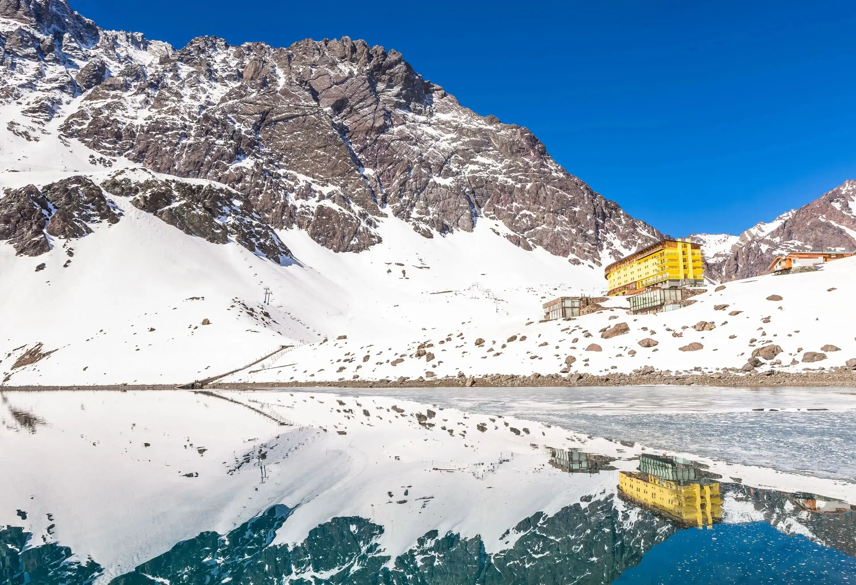 A snow-covered mountain with a yellow structure on its base is reflected on a frozen crystalline lake.