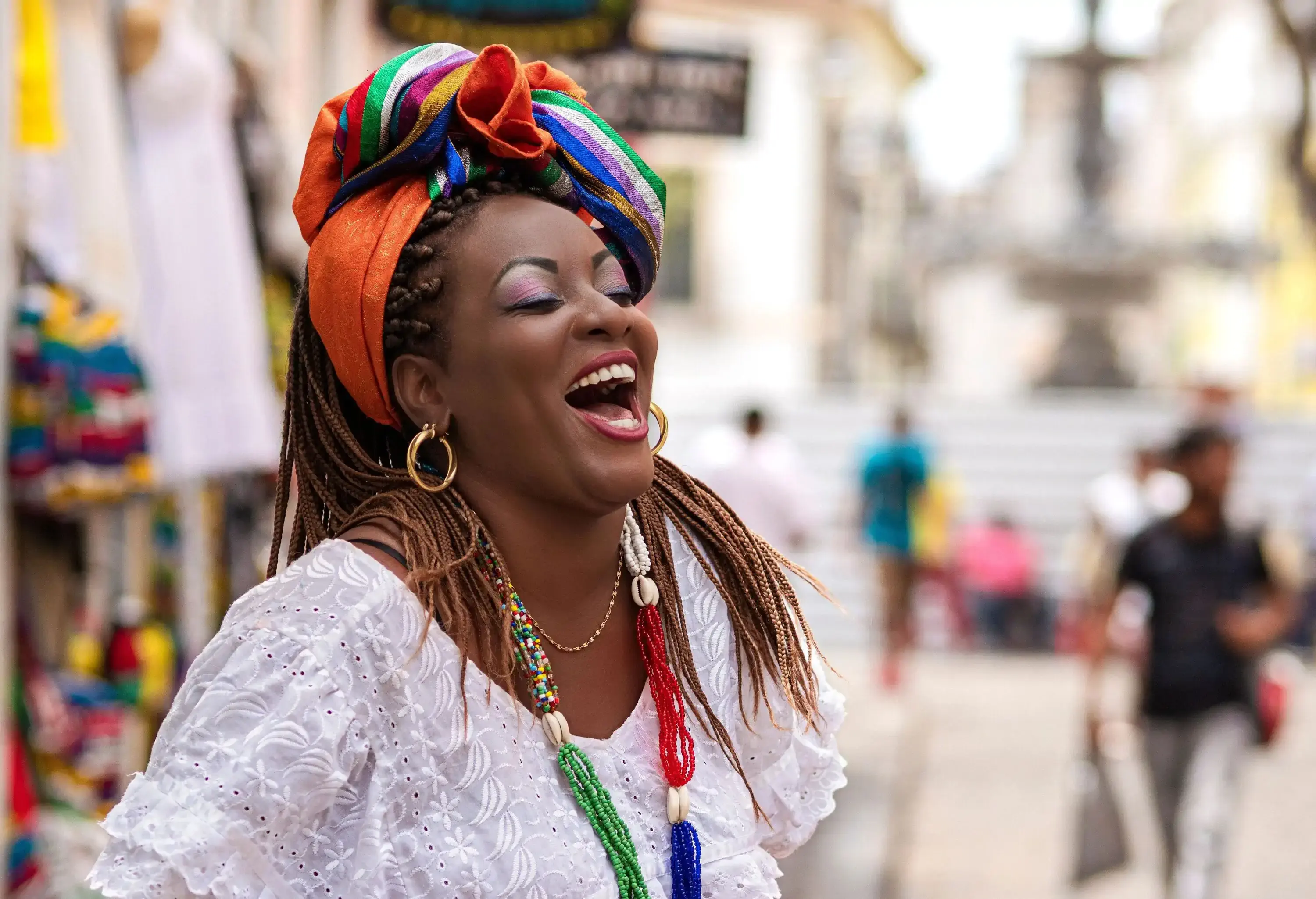 A woman laughing while wearing a white top and a colourful scarf wrapped over her braided hair.