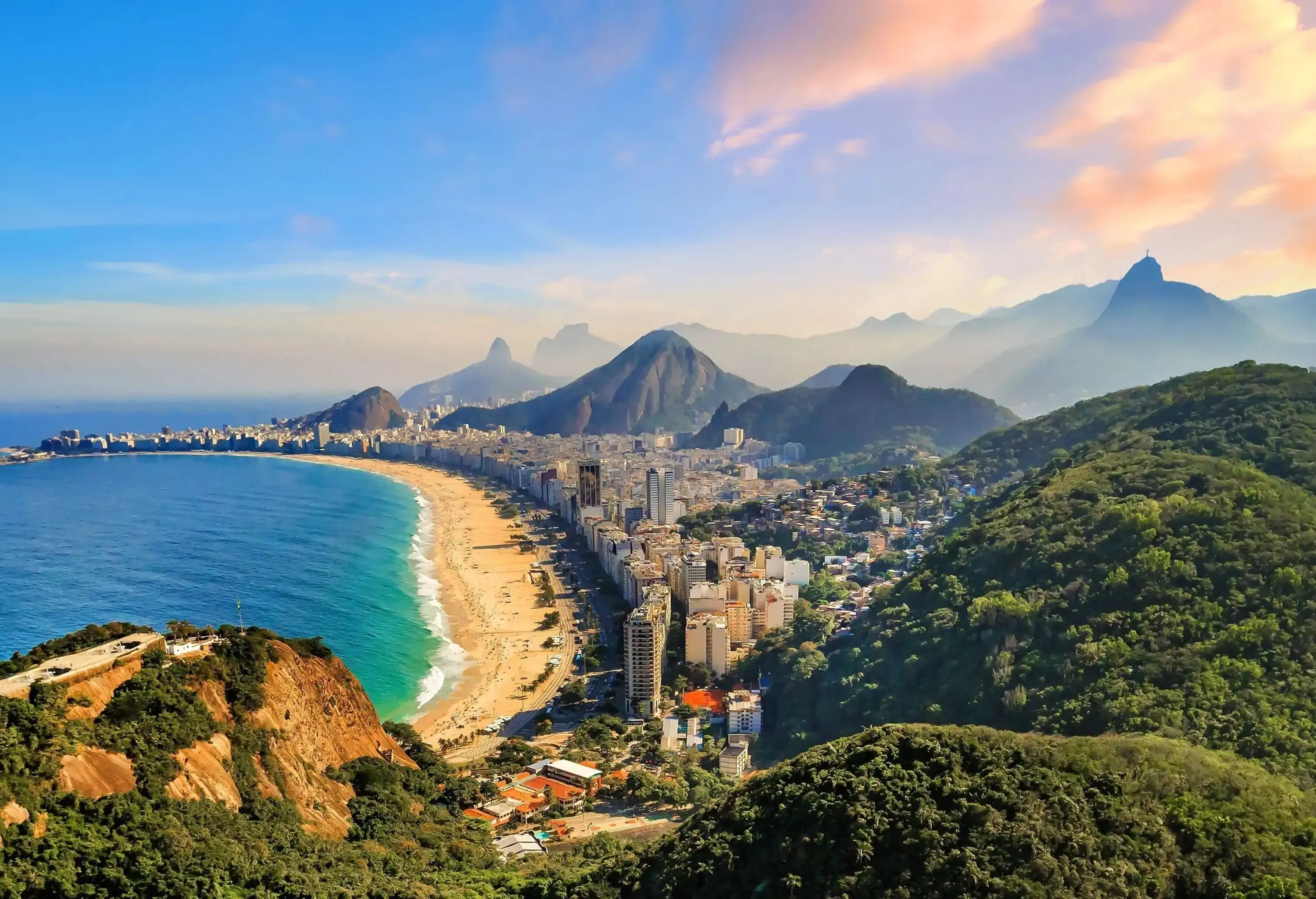 A long and picturesque stretch of beach is bordered by towering buildings and verdant rolling hills.