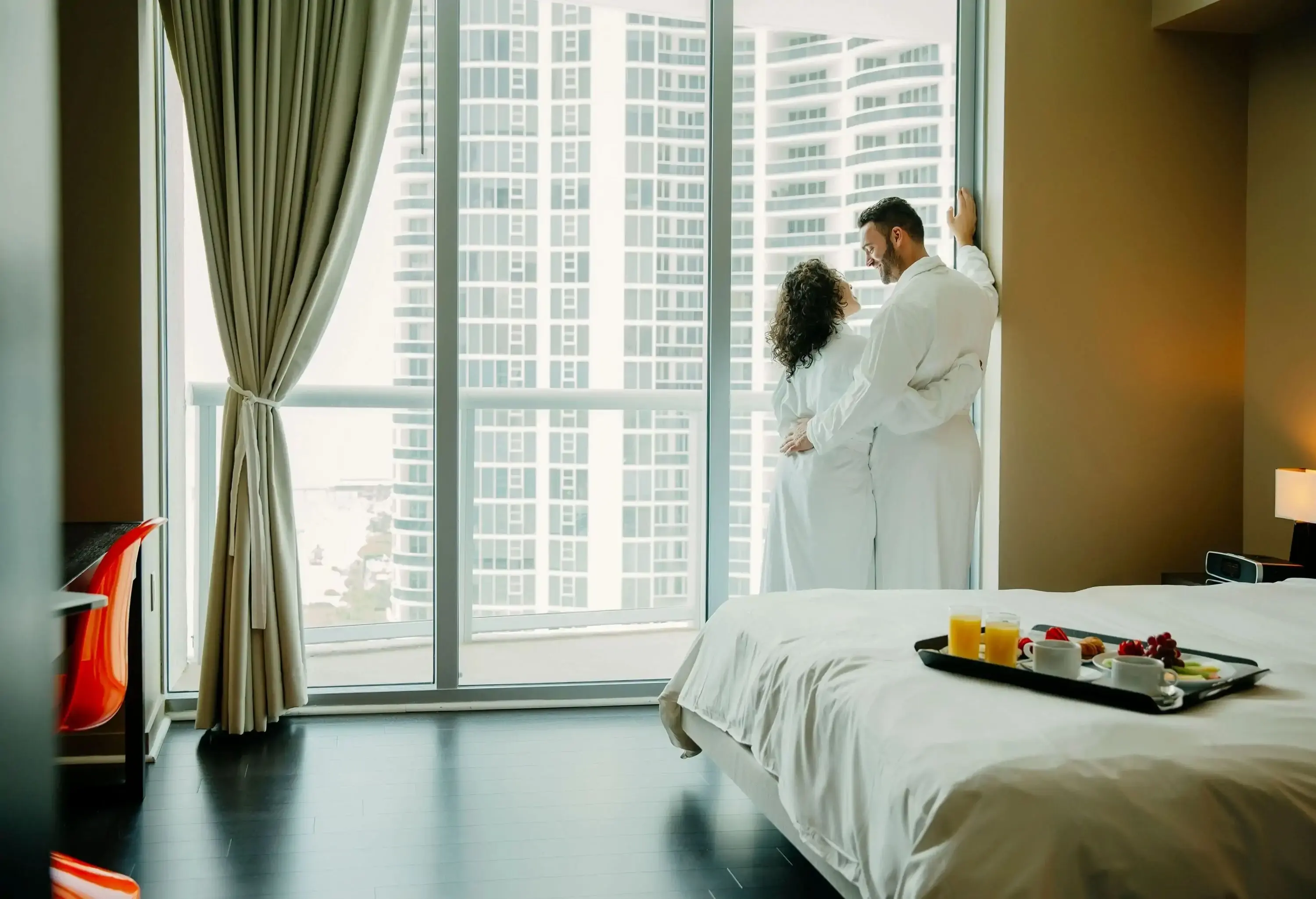 A happy couple in a white robe stands by the glass windows of a luxury hotel bedroom.