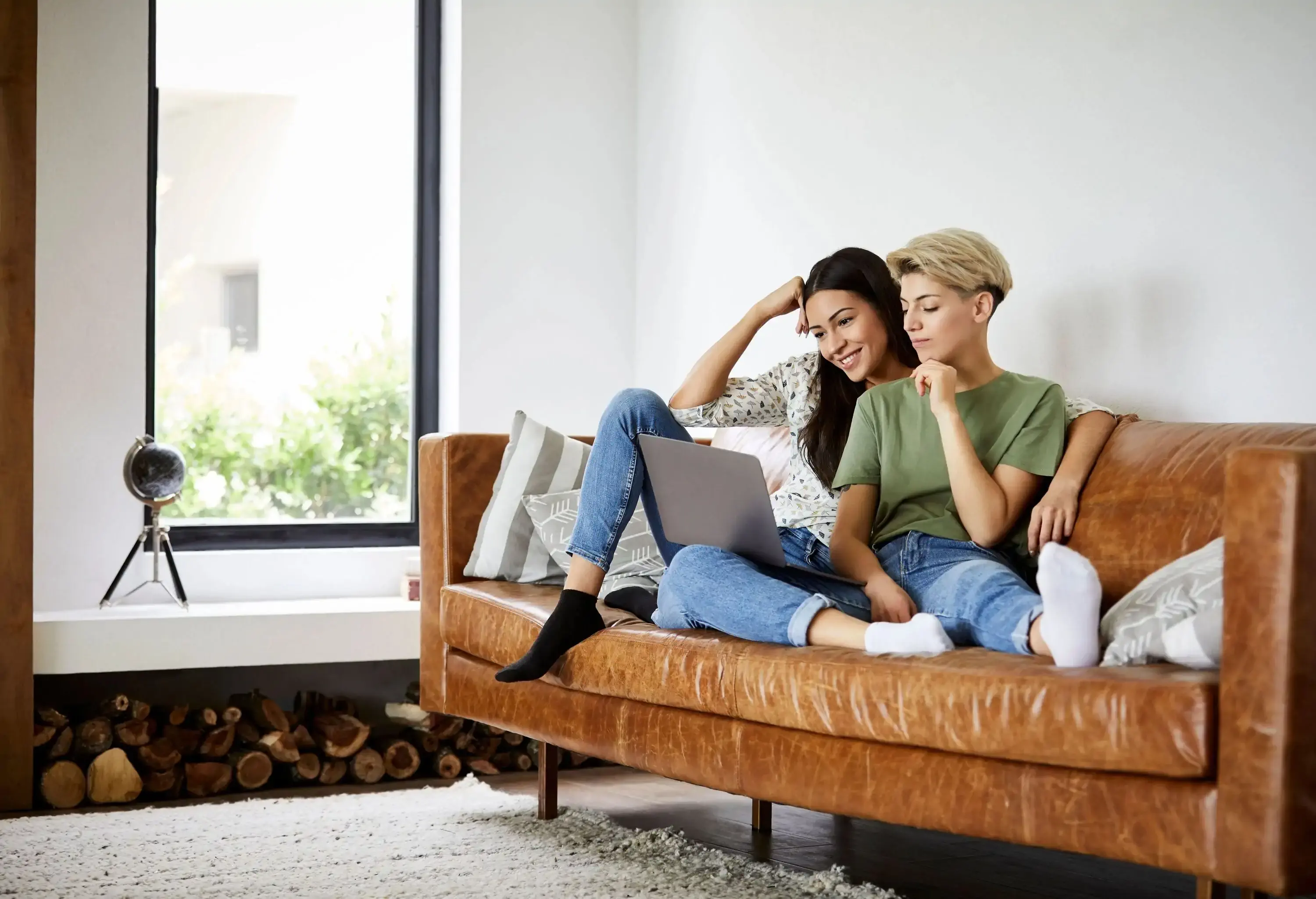 Two women watching on a laptop while sitting on a brown leather couch.
