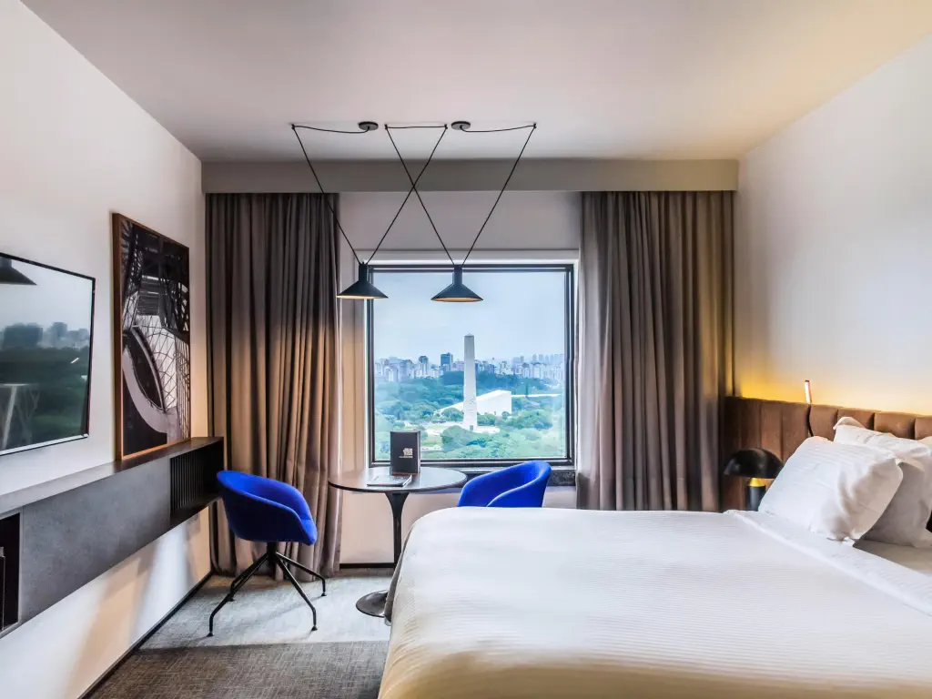 Modern hotel room with a large bed, blue chairs, and a cityscape view through a window. 
