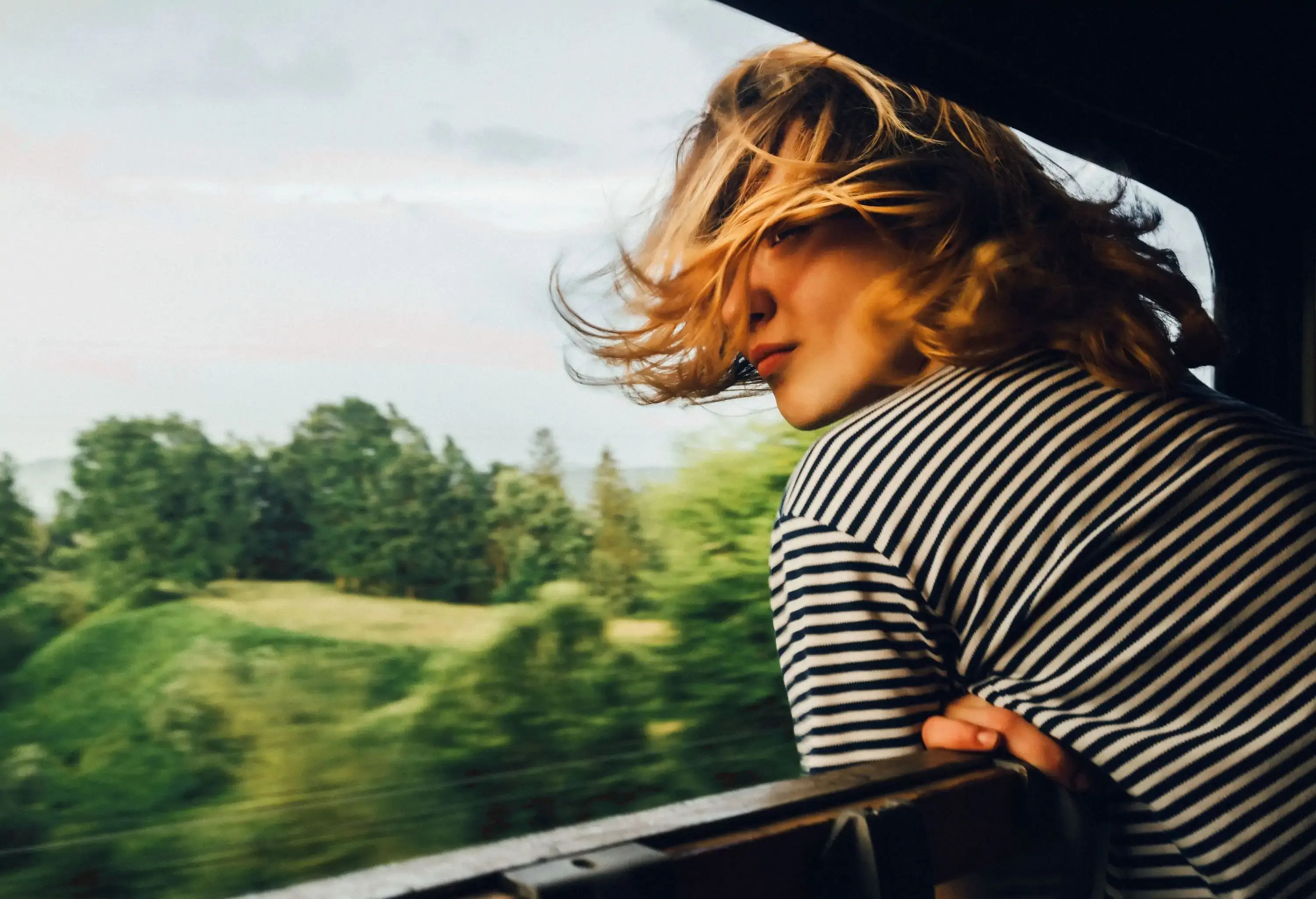 A blonde lady peeking out the window train with messy hair and a view of lush trees.