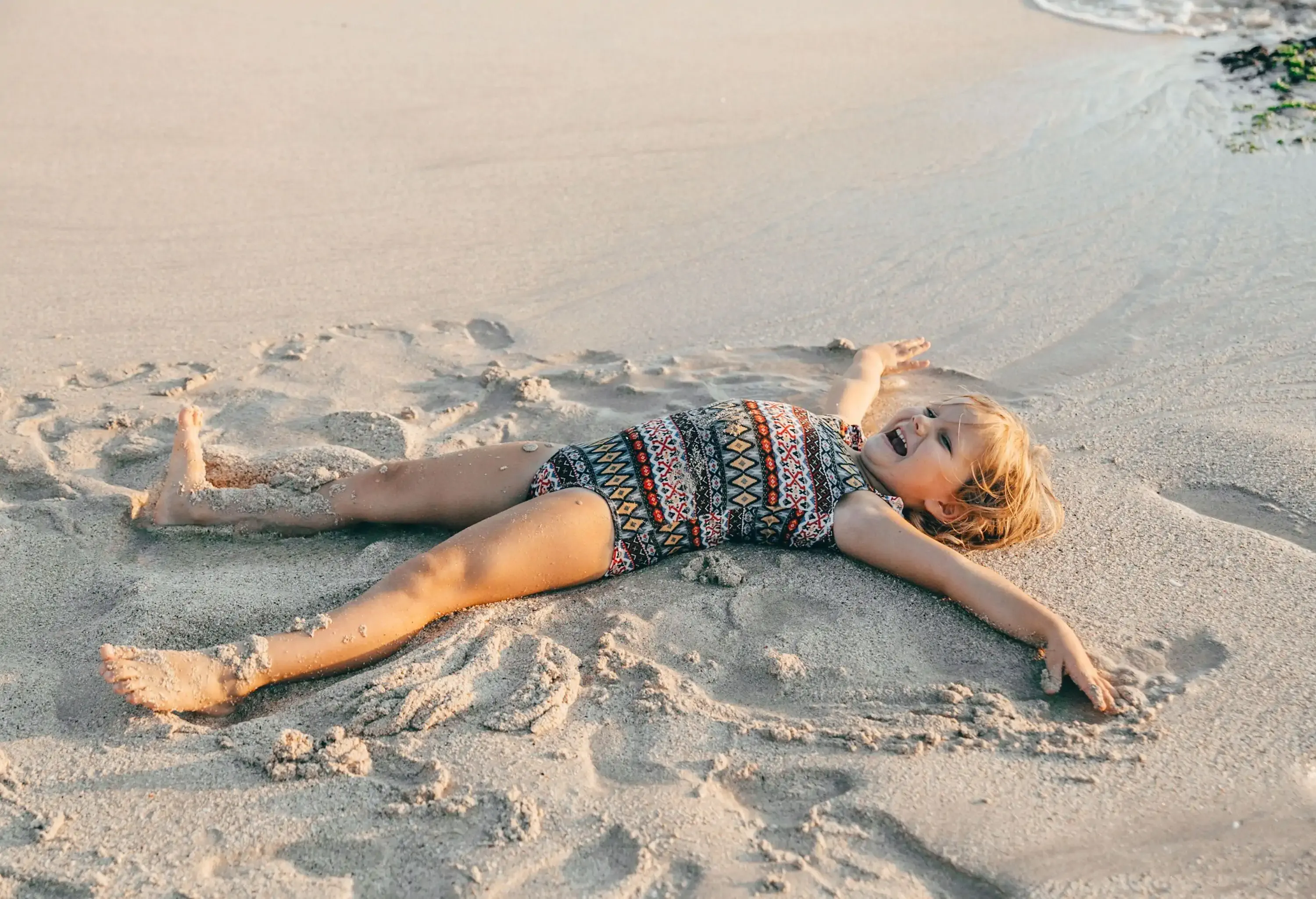 A little girl lying on her back on the sand at the beach, forming a sand angel.