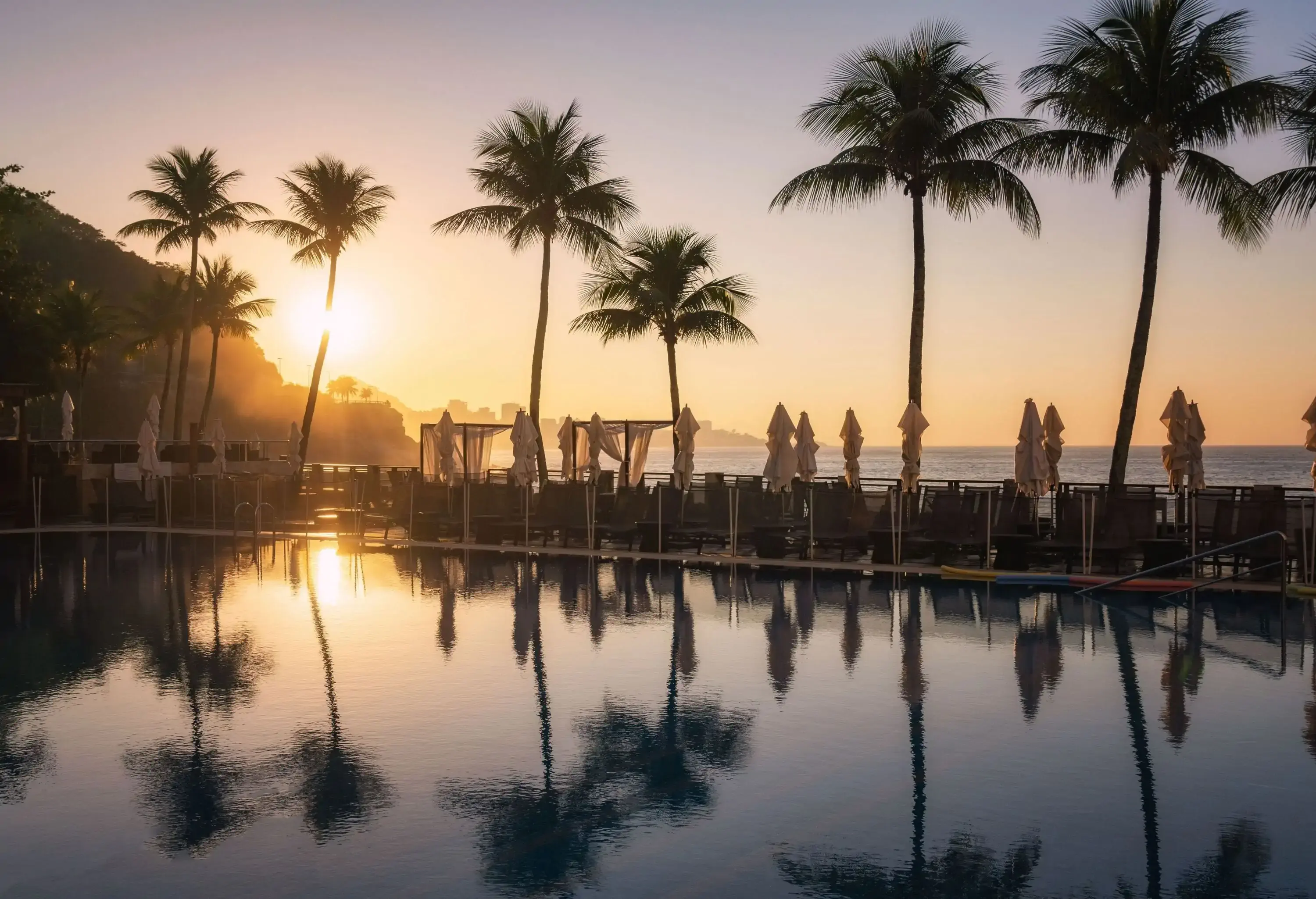 As the sun sets, a serene pool glows with a warm, golden light, surrounded by unoccupied sunbeds and closed parasols, while majestic palm trees line the border.