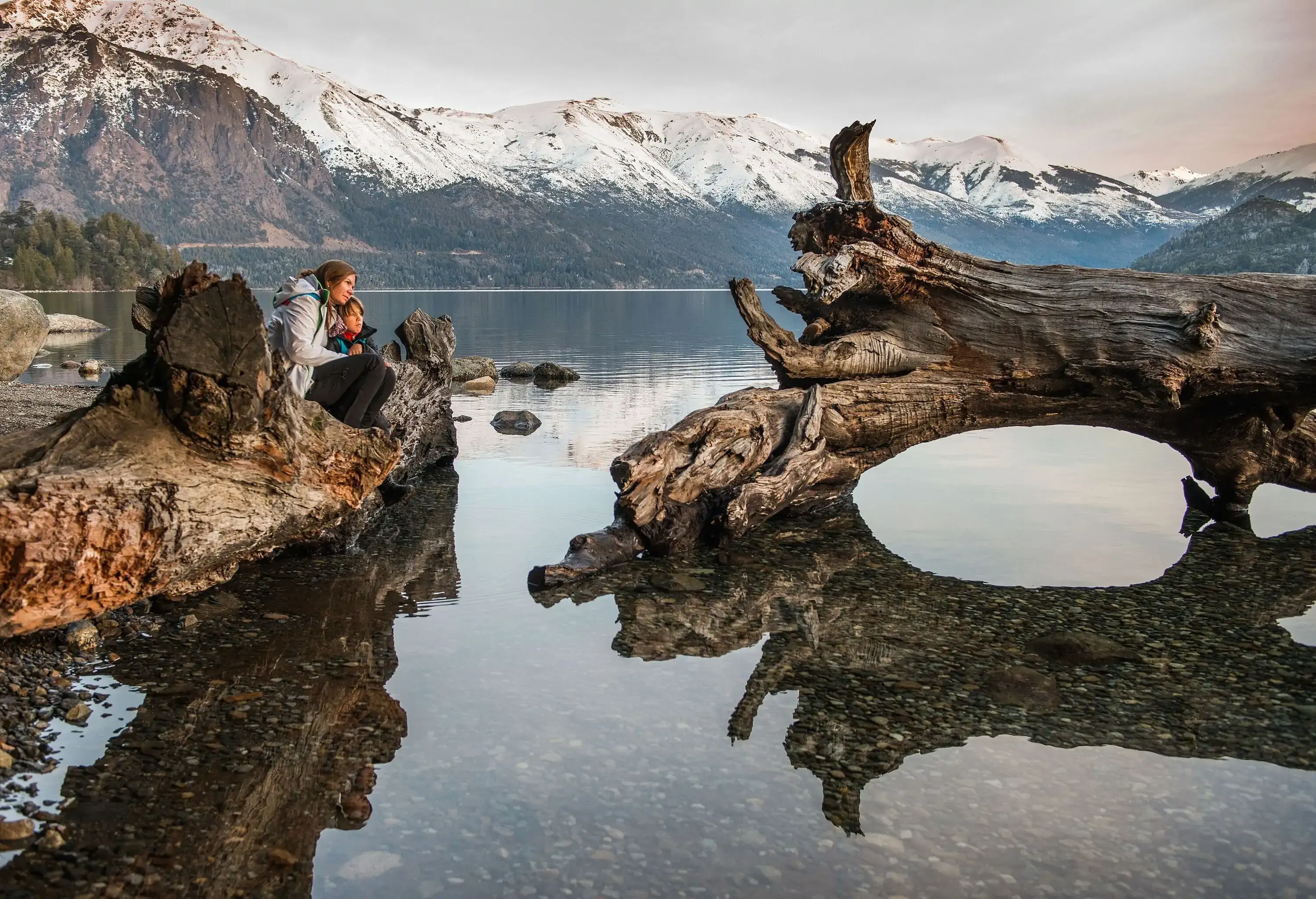 A mother and child share a quiet moment as they sit on a weathered tree trunk by a serene lakeshore, with majestic, snow-dusted mountains forming a breathtaking backdrop in the distance.