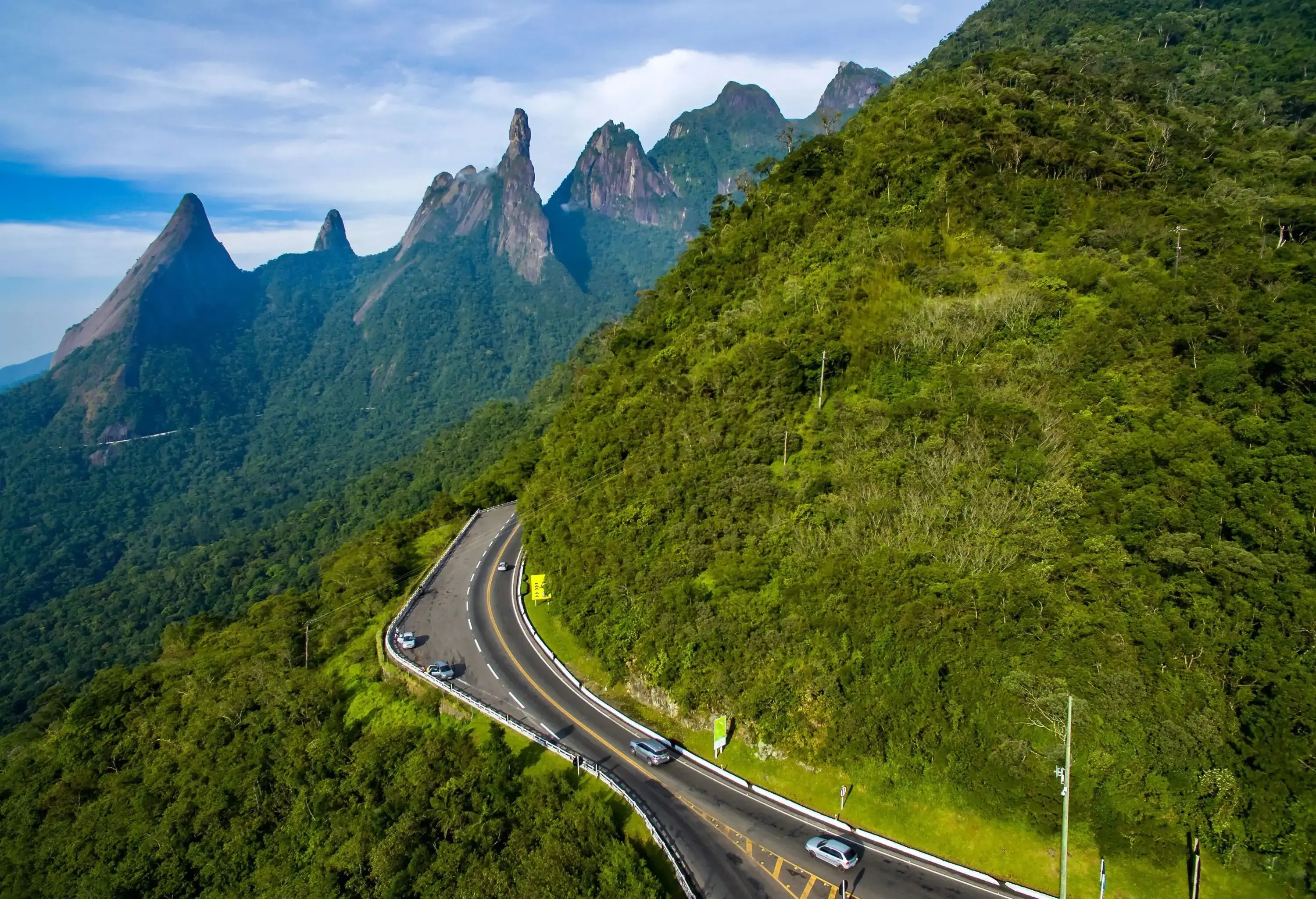 A winding road carves through the lush greenery of the mountainside, with towering rock formations and the majestic Mountain Finger of God providing a breathtaking backdrop.