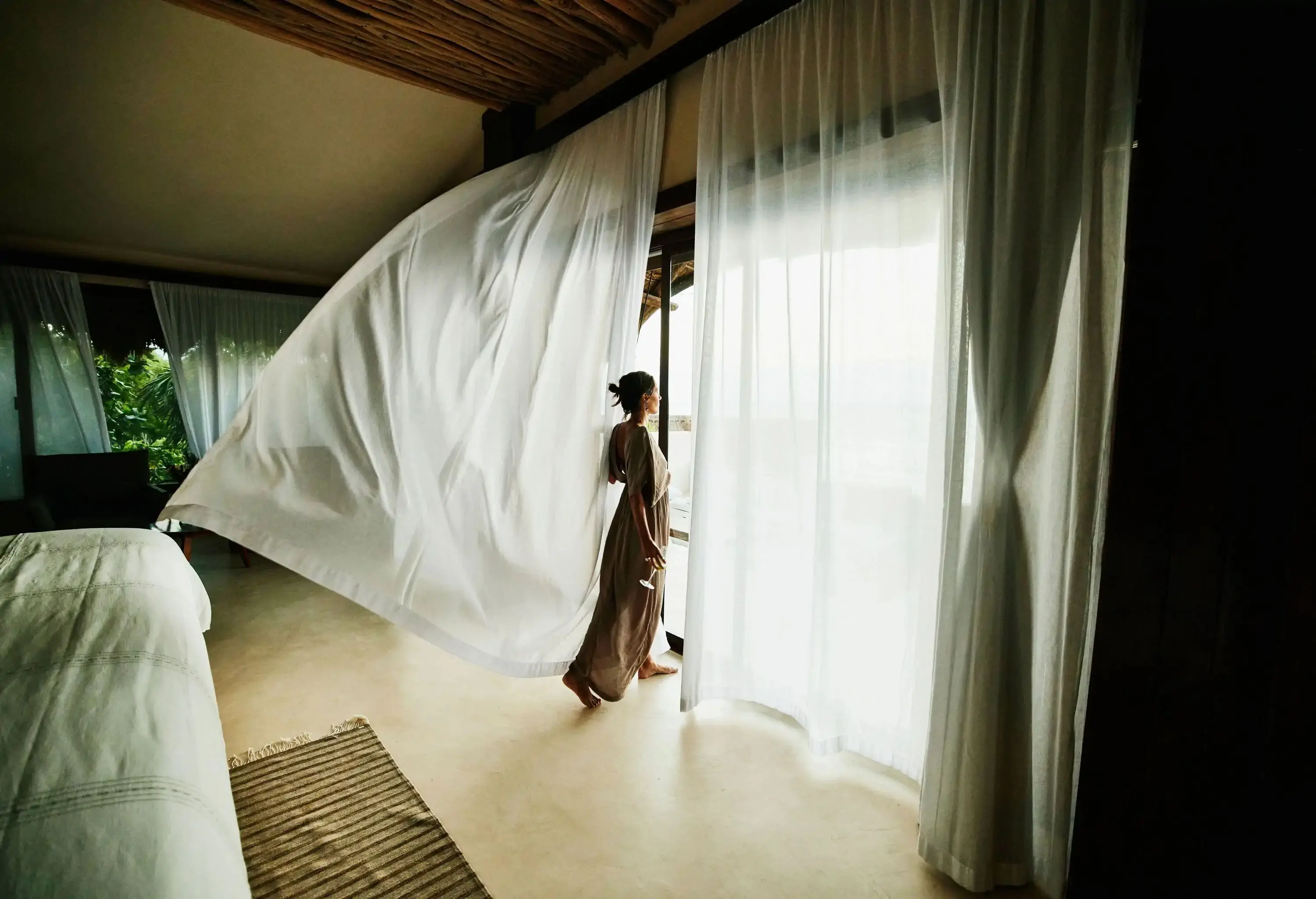 Silouhette of woman looking out of a hotel full window with sea view surrounded by floating curtains