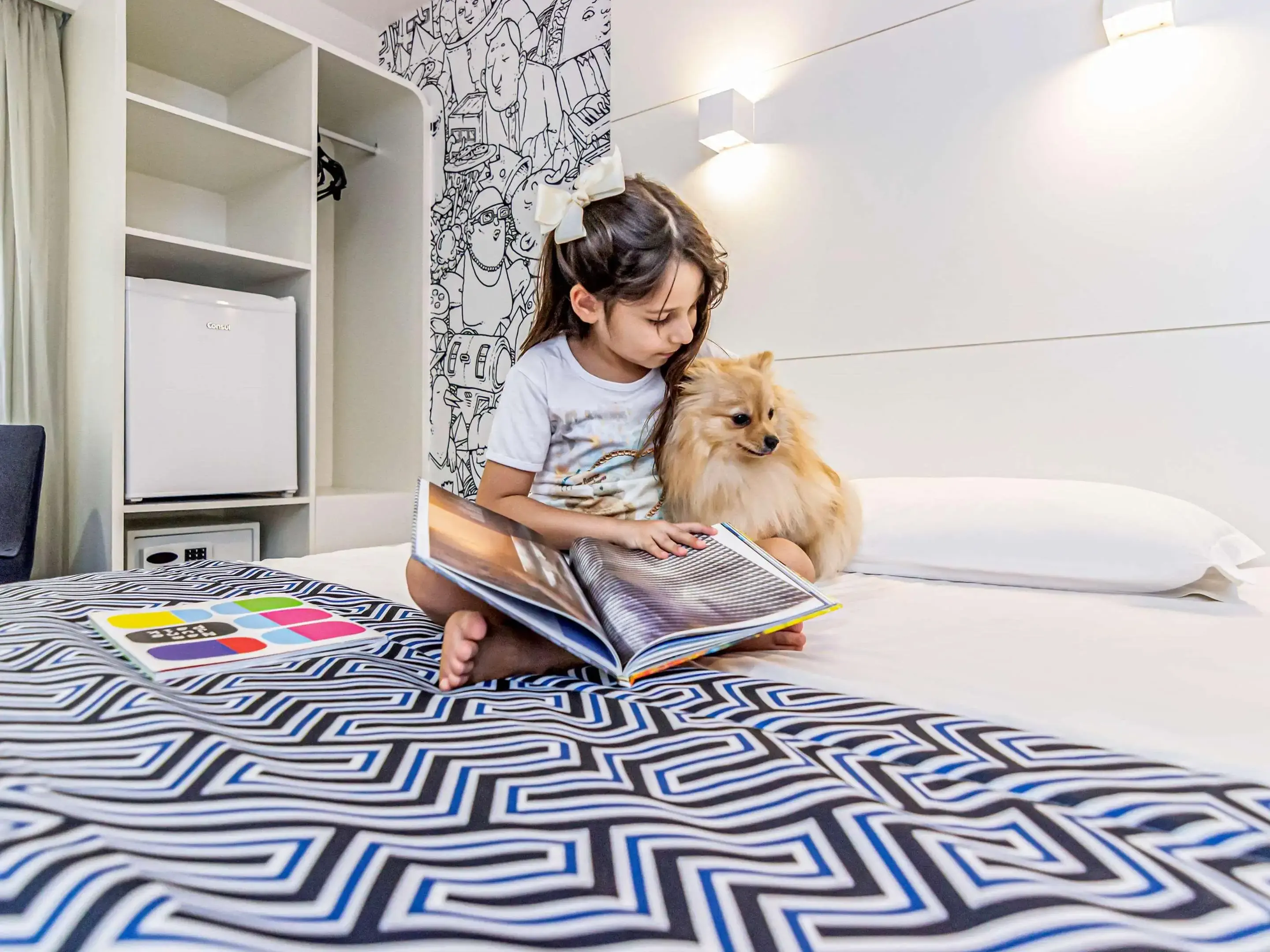 Young girl sitting on a hotel bed reading a book to her pomeranian dog