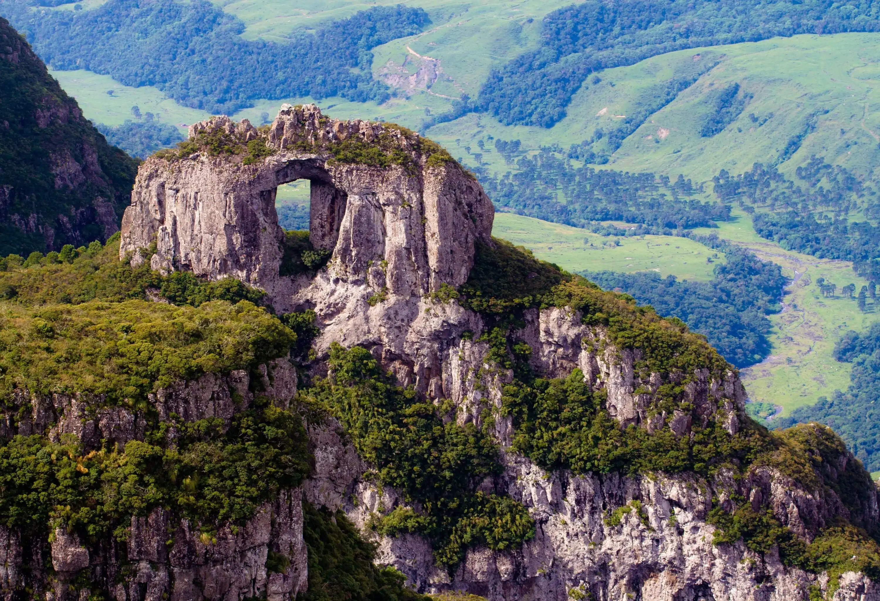 A natural rock formation resembling a gateway, perched atop a rocky mountain and set against the backdrop of a lush green, sloping landscape.