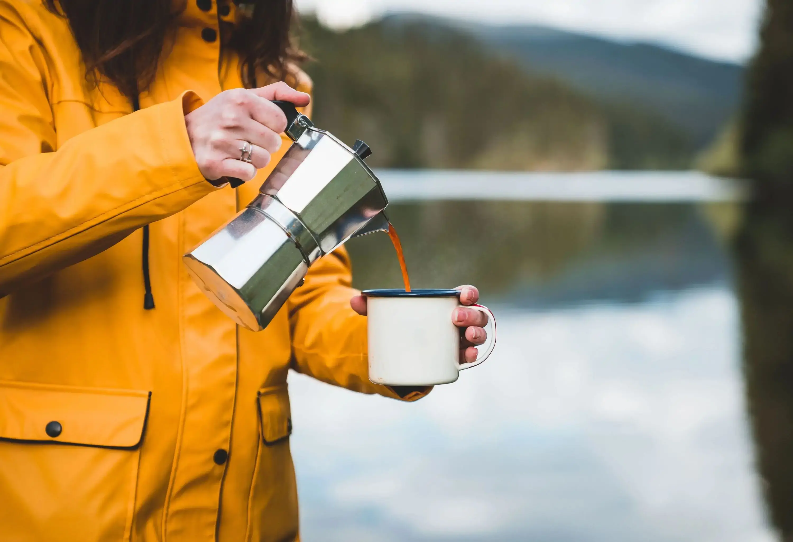 A woman wearing a yellow coat pouring coffee into a cup from a moka pot.