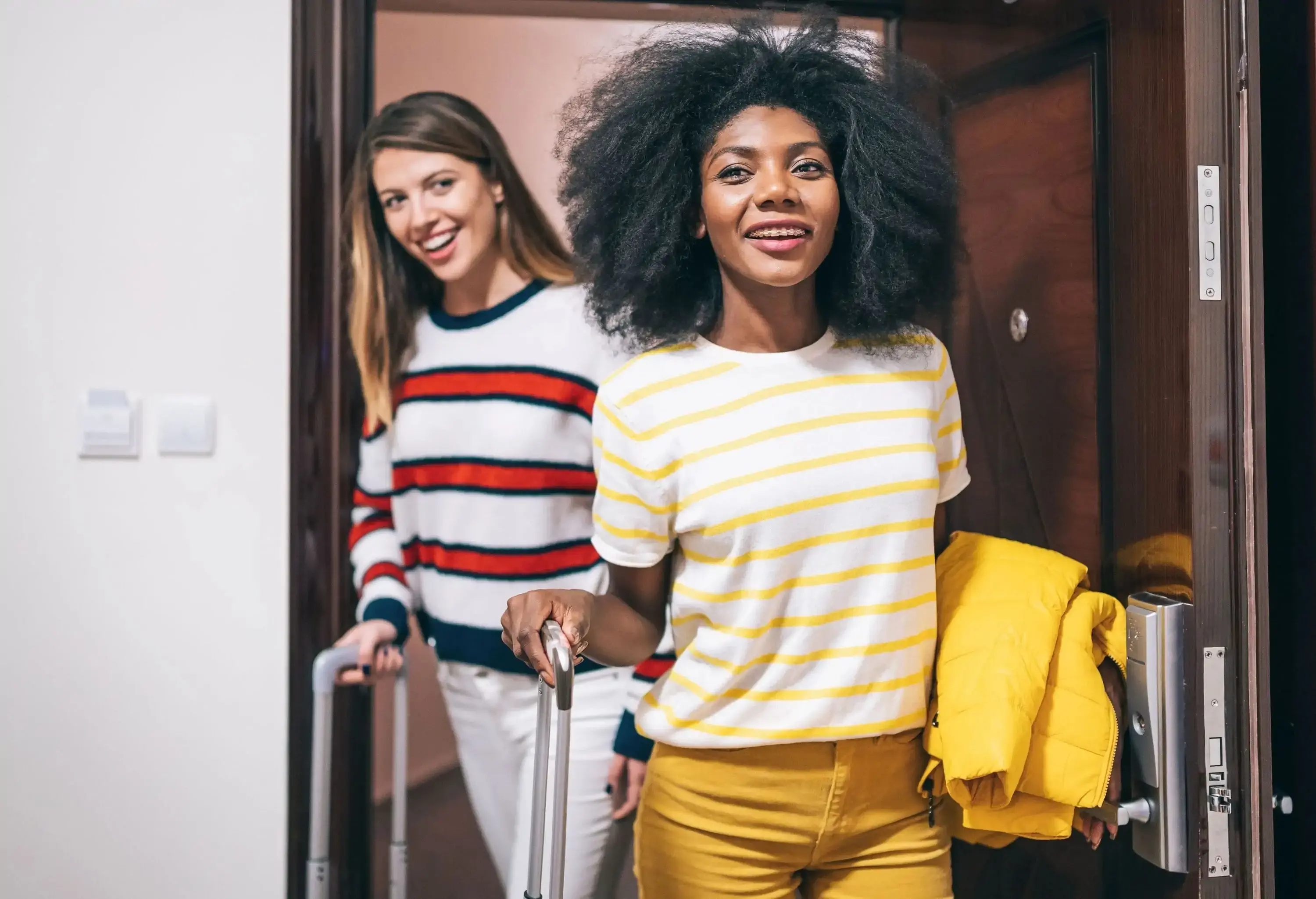 Female friends, both wearing striped shirts and beaming with excitement, arrive at their hotel and open their room door while effortlessly sliding their wheeled luggage, ready for a memorable stay.