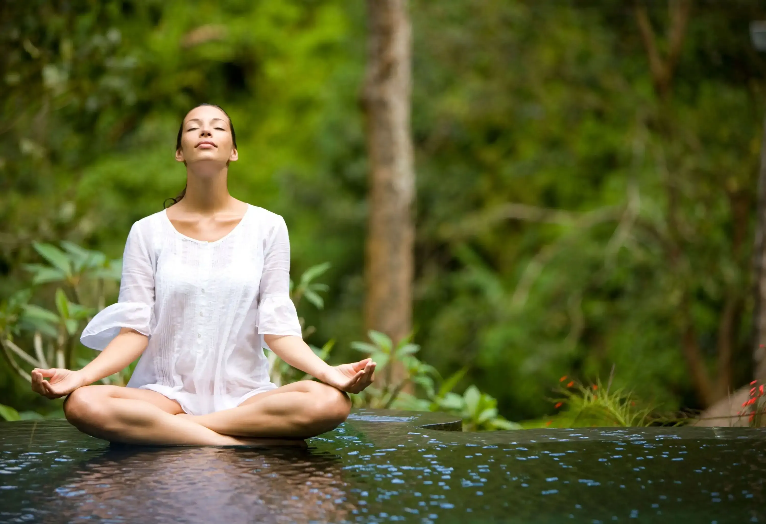 A woman with her eyes closed doing yoga at the edge of an infinity pool amidst the lush plants.