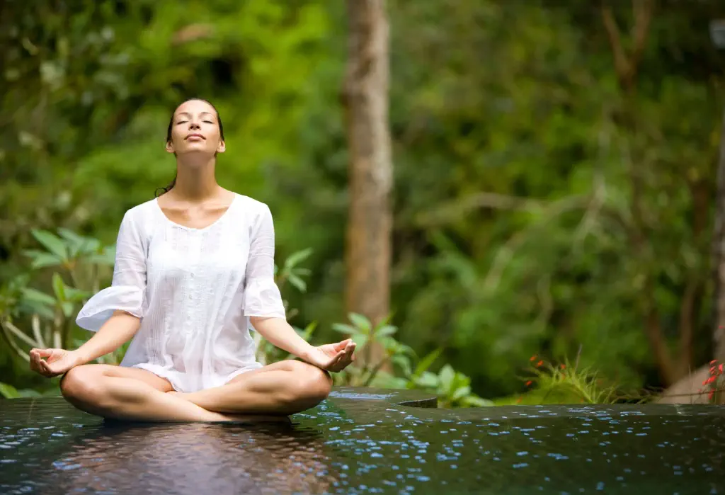 A woman with her eyes closed doing yoga at the edge of an infinity pool amidst the lush plants.