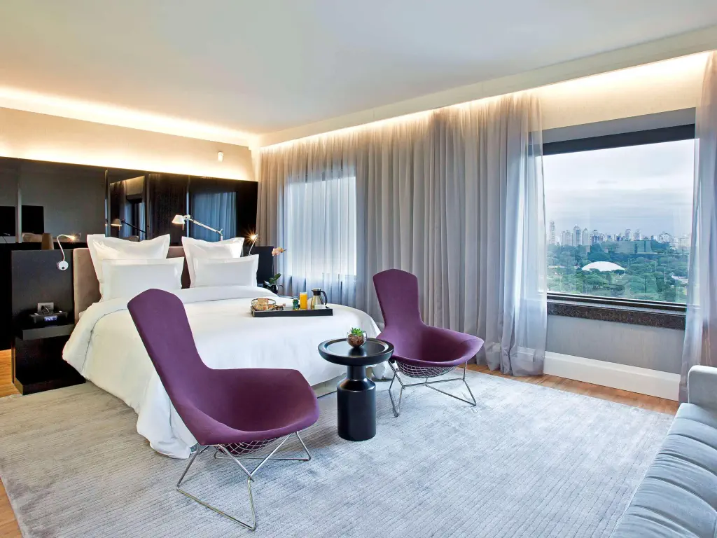Modern hotel room with a large bed, two purple chairs, a small round table, floor-to-ceiling windows with city views, and light-colored furnishings.
