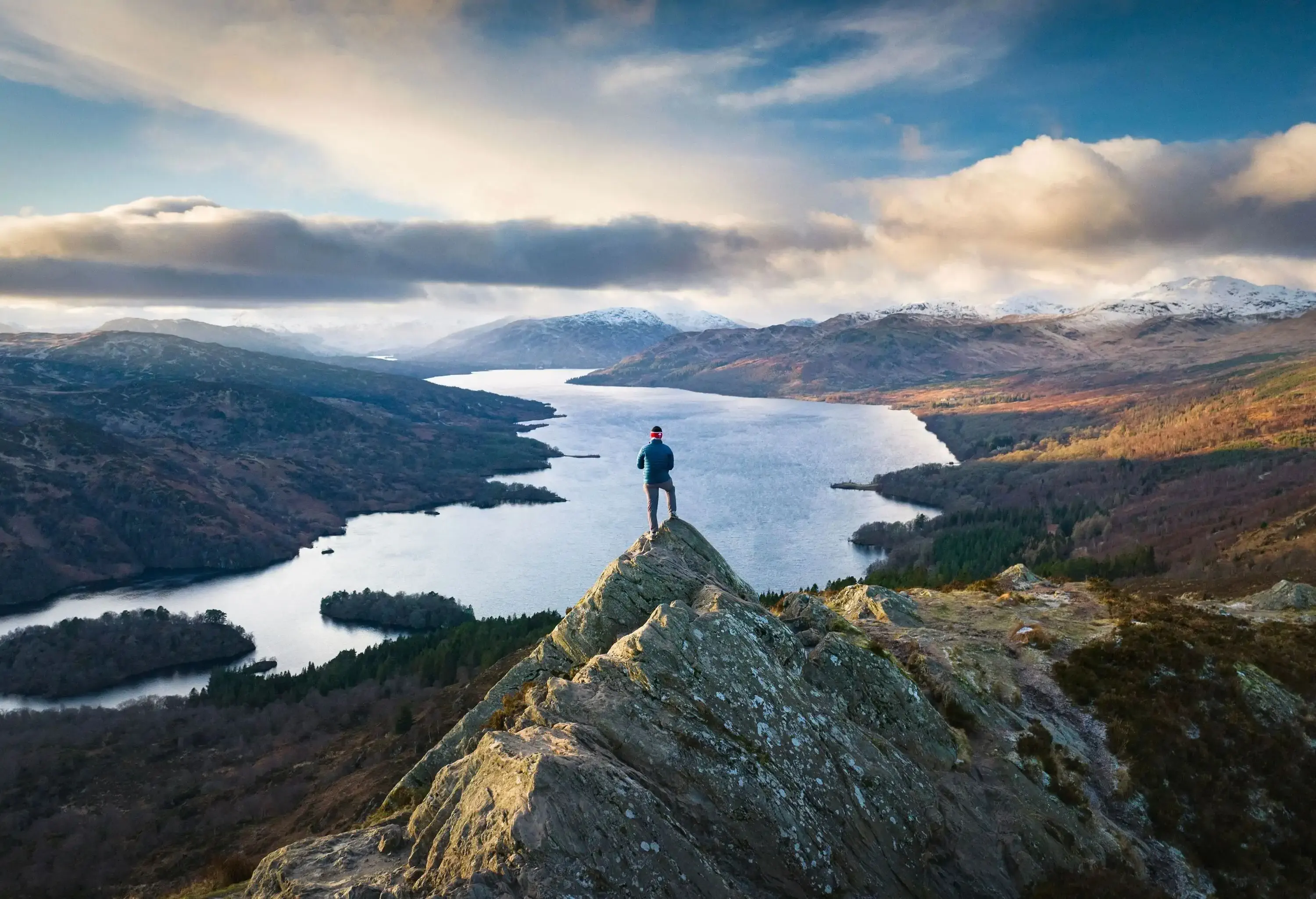A person stands on a rocky peak overlooking a long river winding through a valley surrounded by hills and distant mountains beneath a cloudy sky.