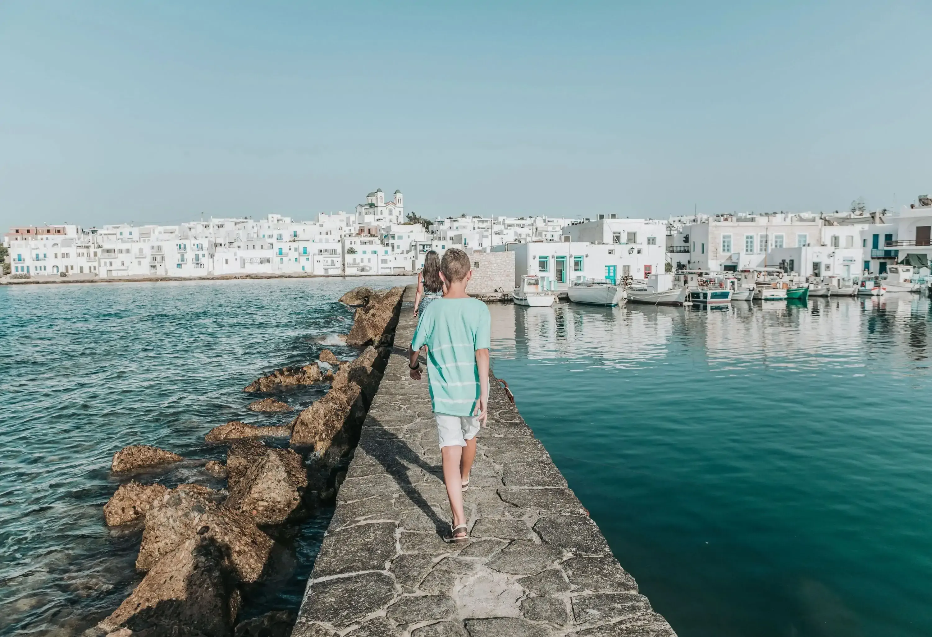 A young boy and girl walking on a stone footbridge across the sea with a white village on the shore.