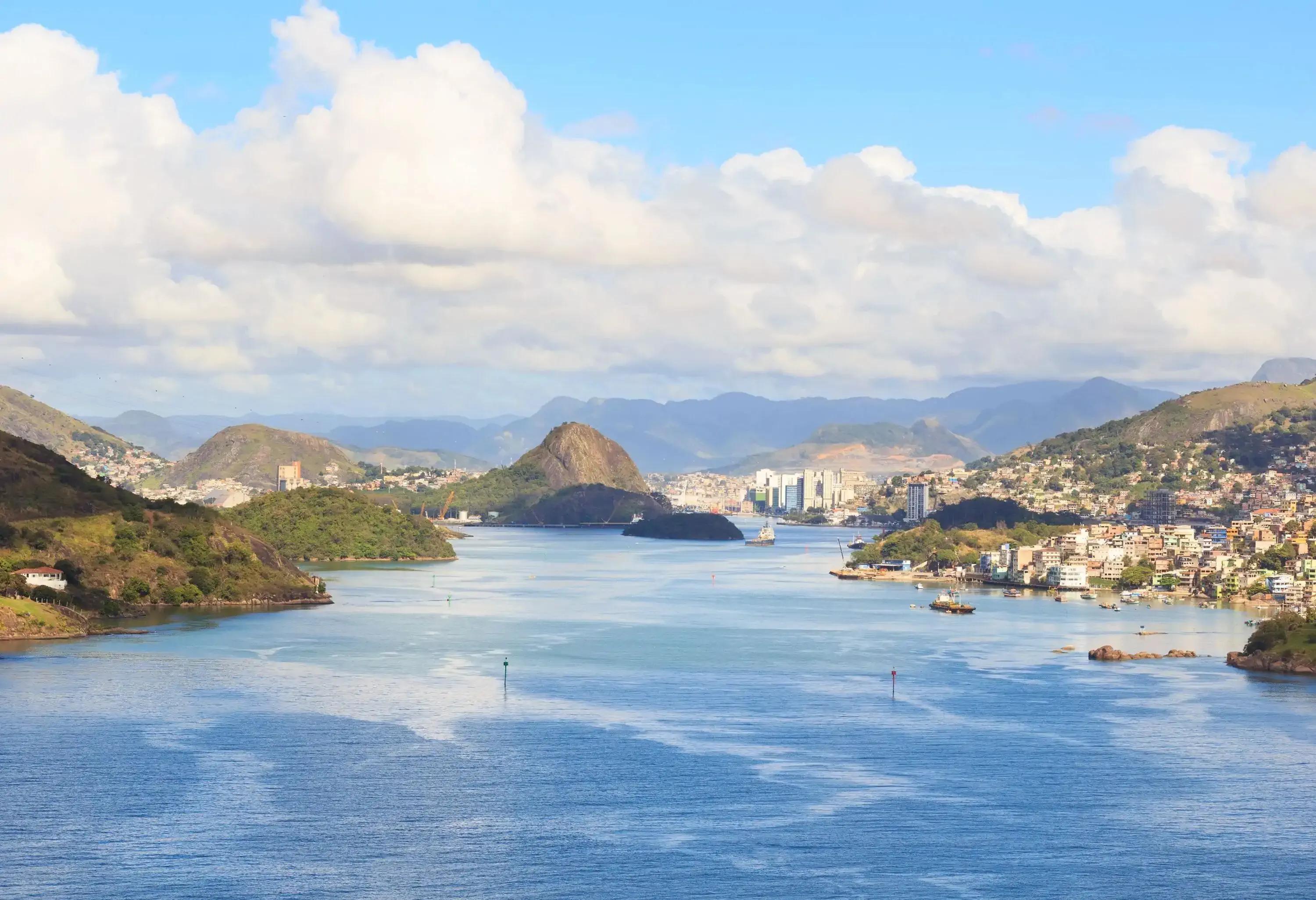 A tranquil sea surrounded by lush hilly islands with houses and buildings captured under the cloudy blue sky.