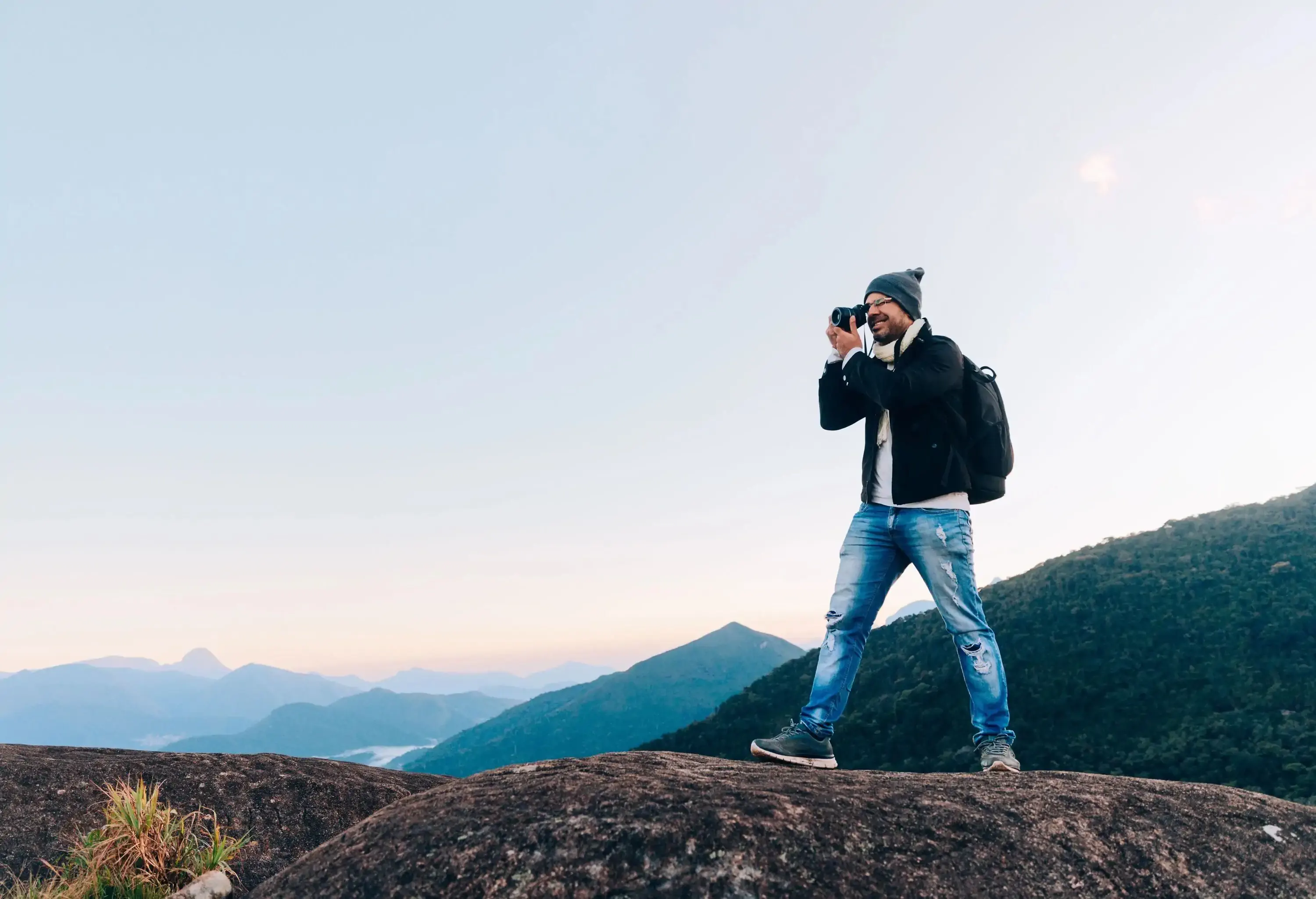 A photographer stands atop a large rock, capturing the beauty of the outdoors as verdant rolling hills unfold behind him, gradually fading into the distance and blending with the horizon.
