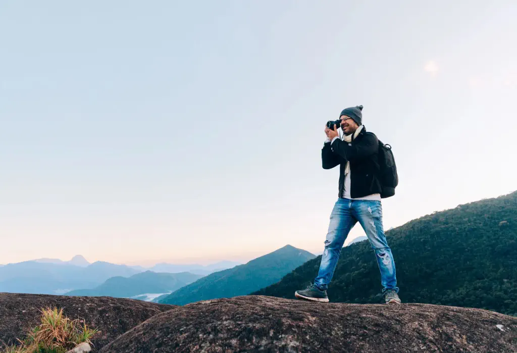 A photographer stands atop a large rock, capturing the beauty of the outdoors as verdant rolling hills unfold behind him, gradually fading into the distance and blending with the horizon.