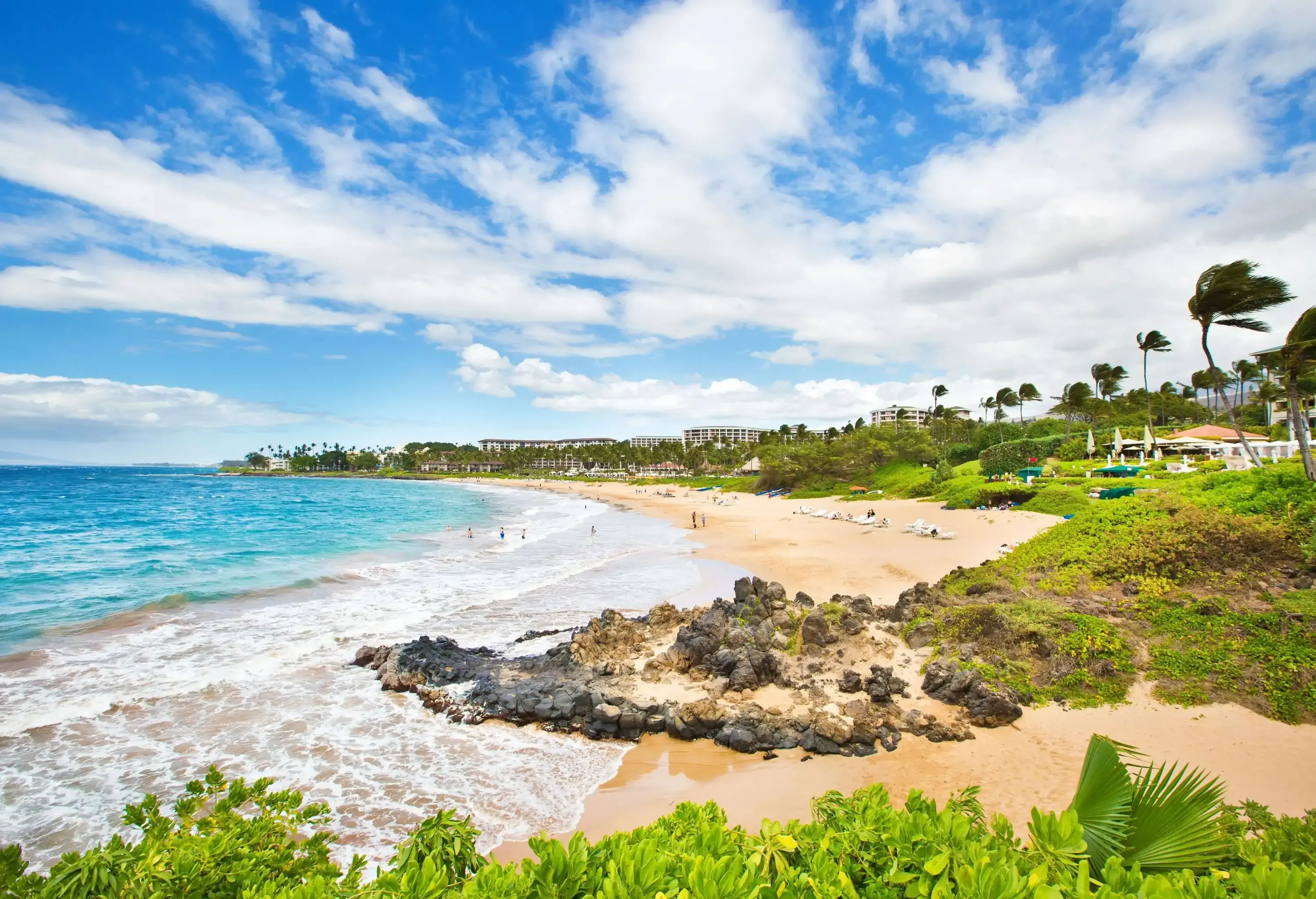 People swim on a beach with mild waves and rocky coastlines along a row of resort and hotel buildings.