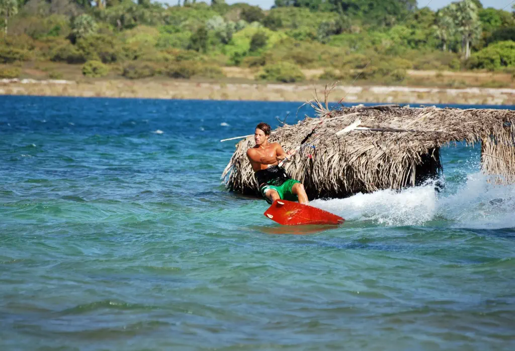 A man kitesurfing in the ocean waves beside a submerged thatched cottage.