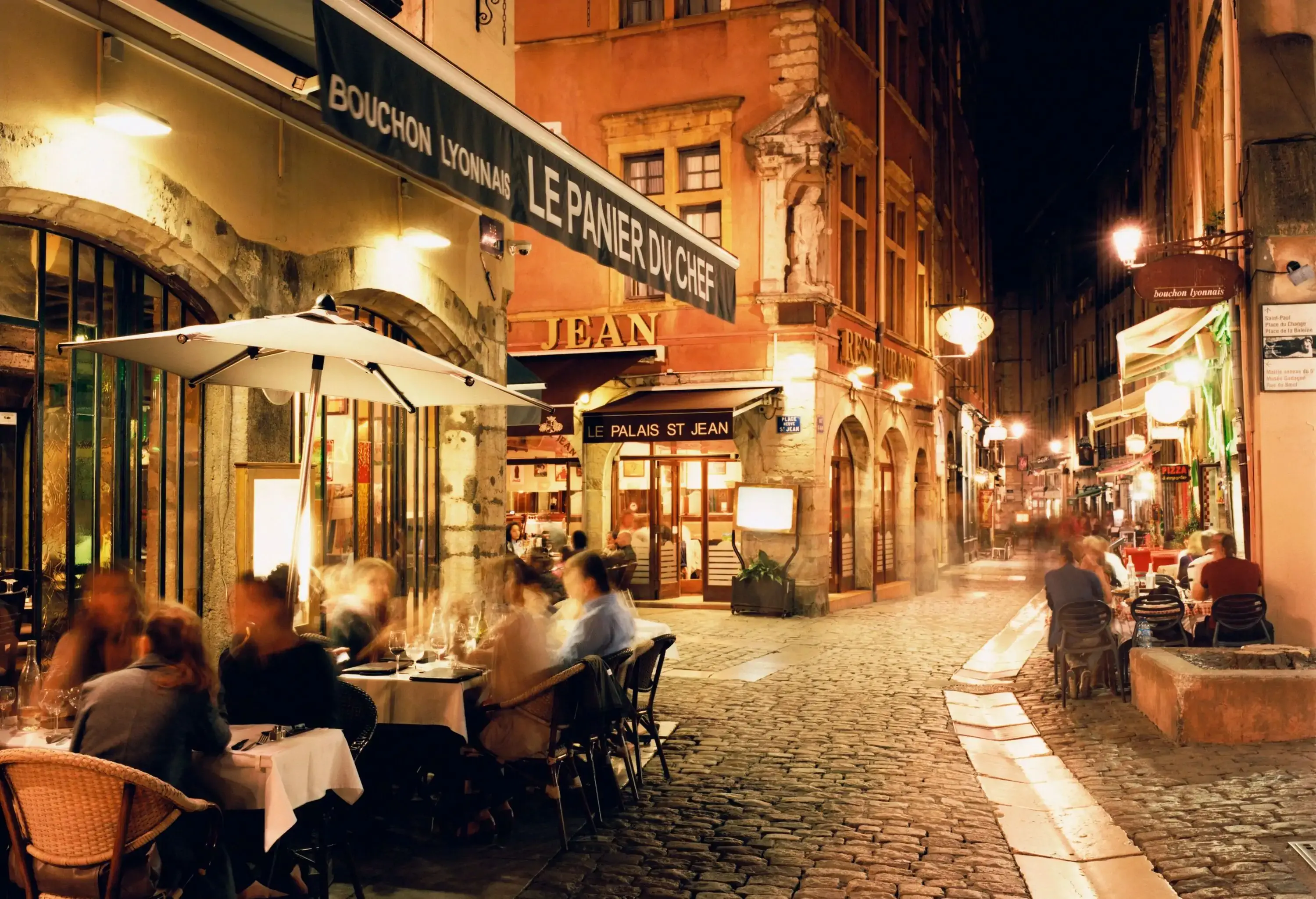 Customers enjoy outdoor dining along a narrow cobbled street in the middle of buildings.