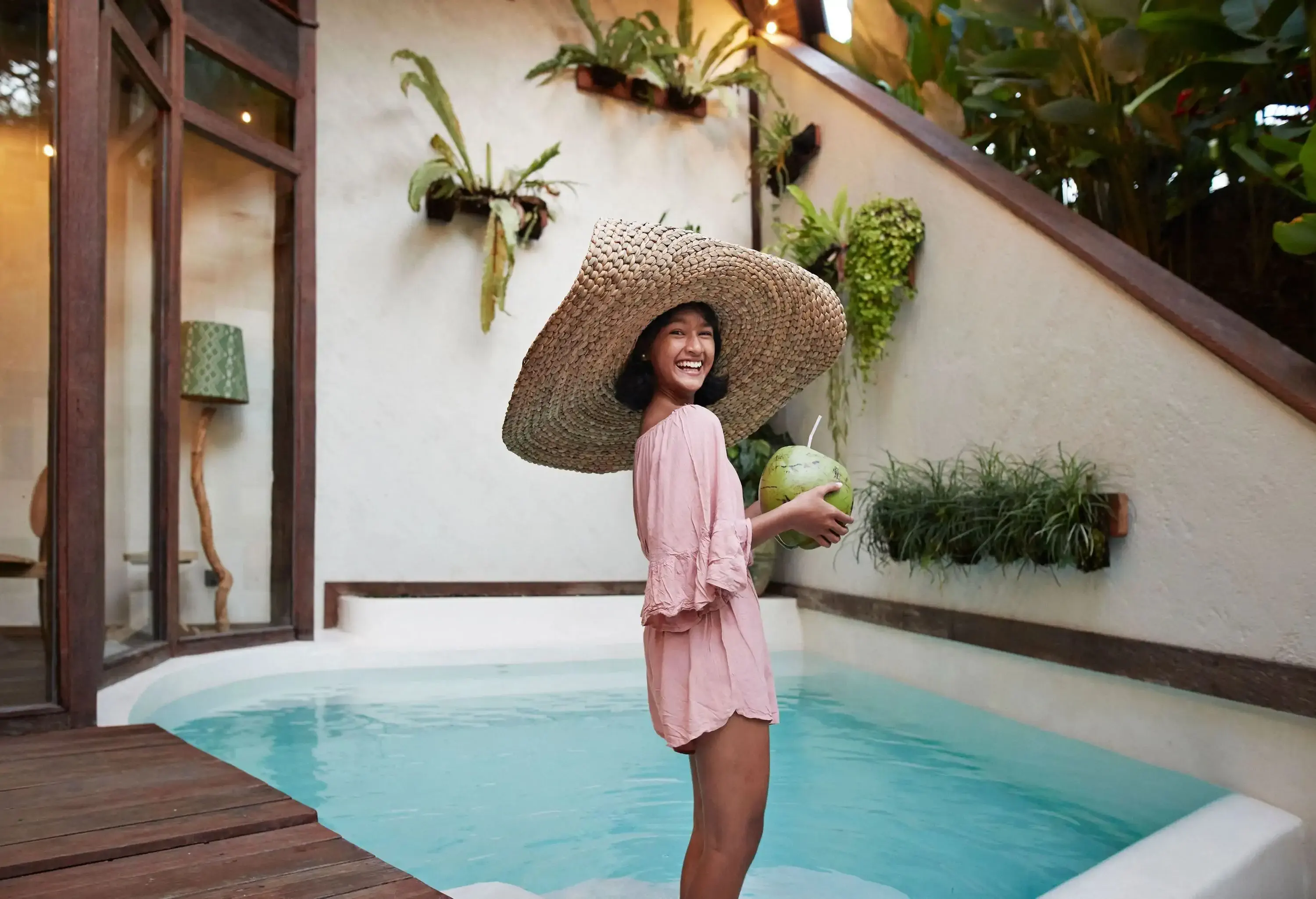 A young woman in a large straw hat smiles while holding a coconut drink in a plunge pool.